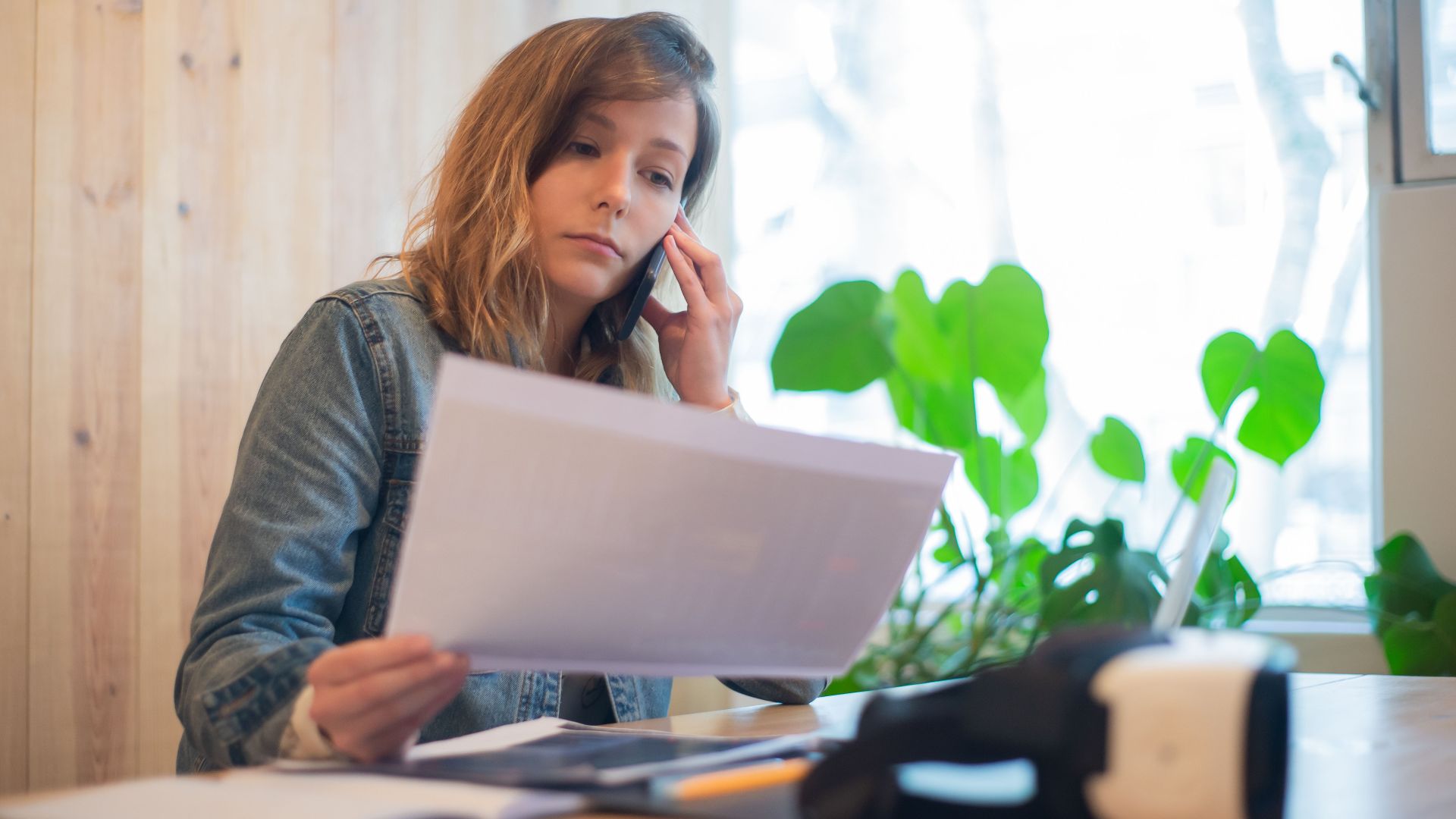 Businesswoman reviewing paperwork while on a call at a modern office workspace.