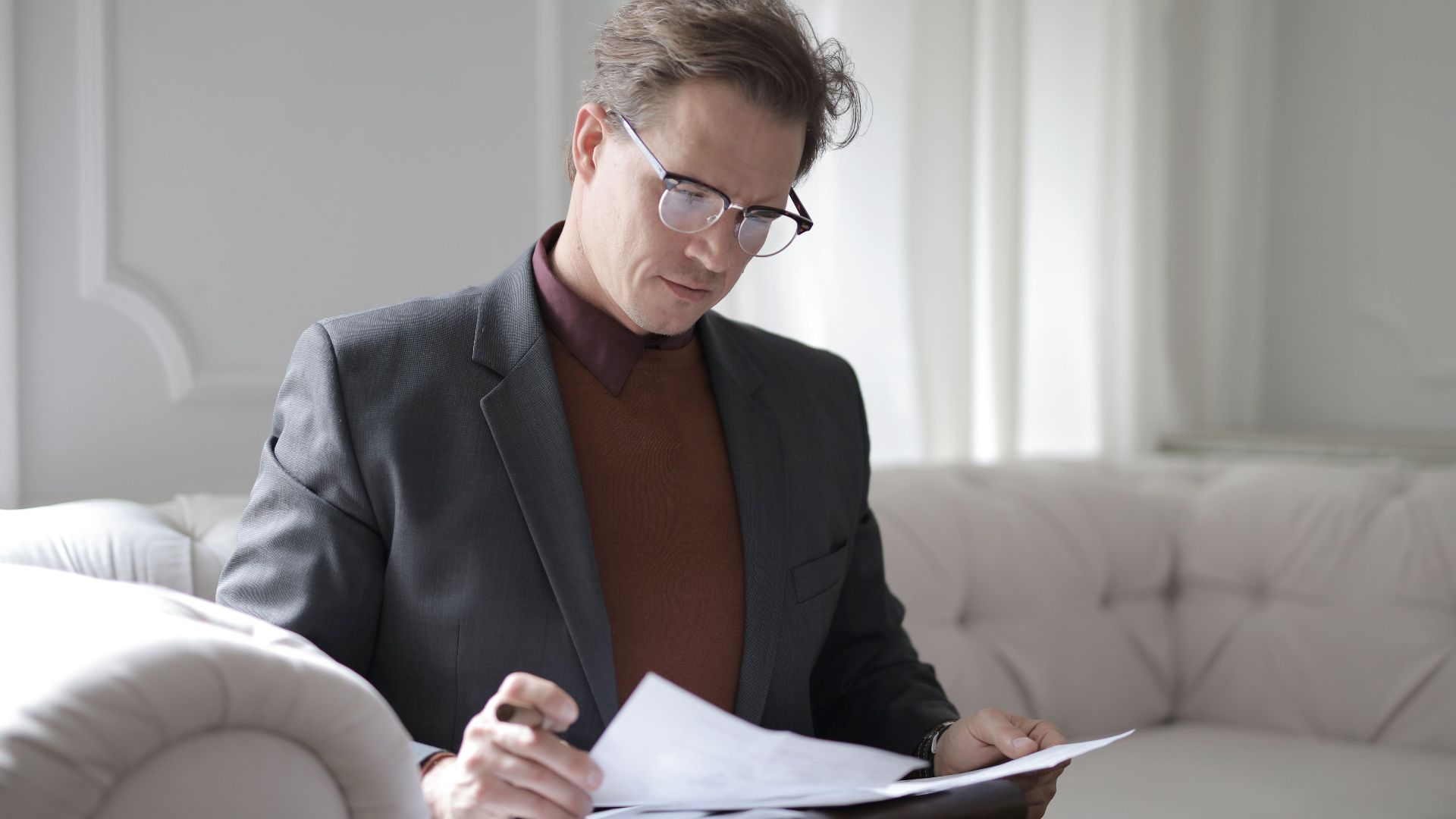 Elegant adult man in jacket and glasses looking through documents while sitting on white sofa in luxury room