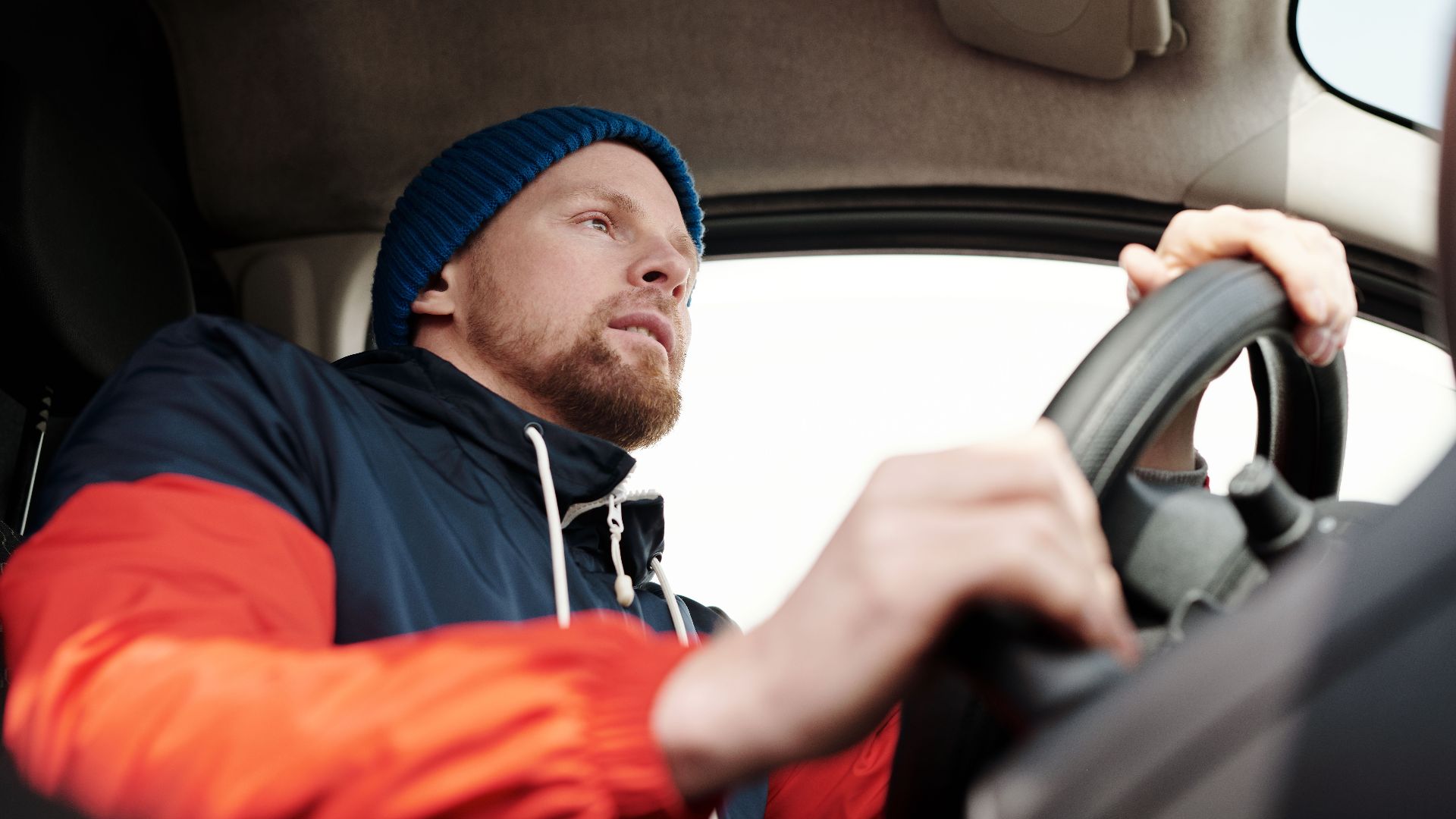 Low angle shot of a bearded man driving a car, wearing a hoodie and knit cap.
