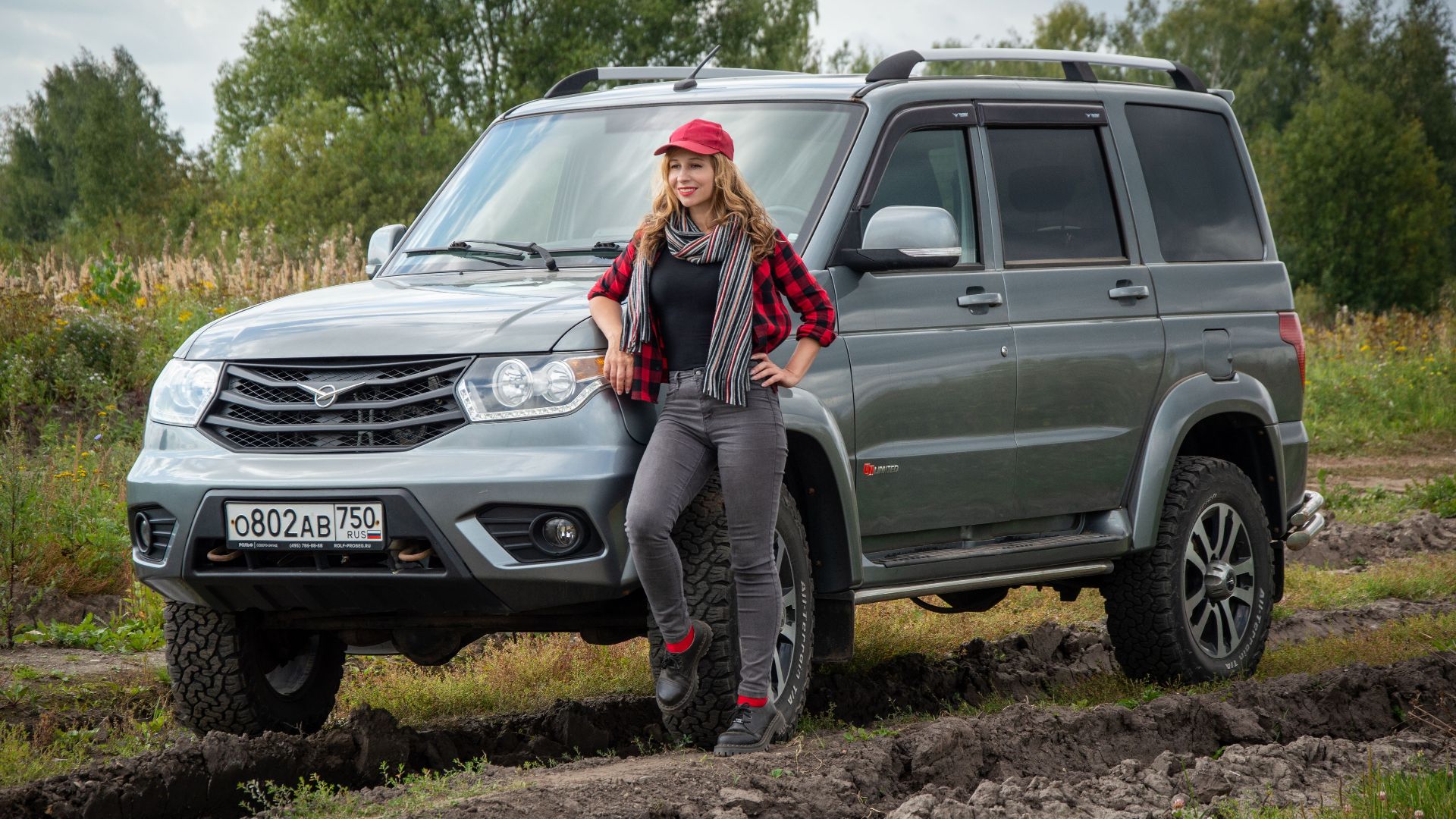 Young woman in casual attire posing confidently with a silver SUV on a rural dirt road, surrounded by greenery.