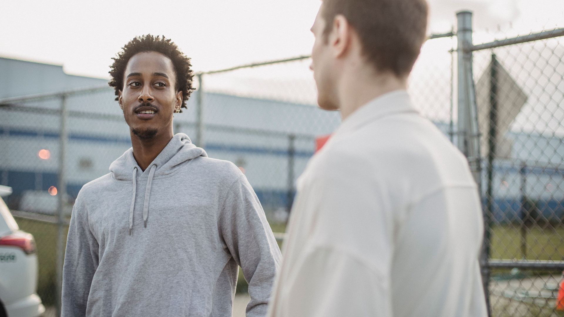 African American male standing with friend against metal grid near car on parking lot
