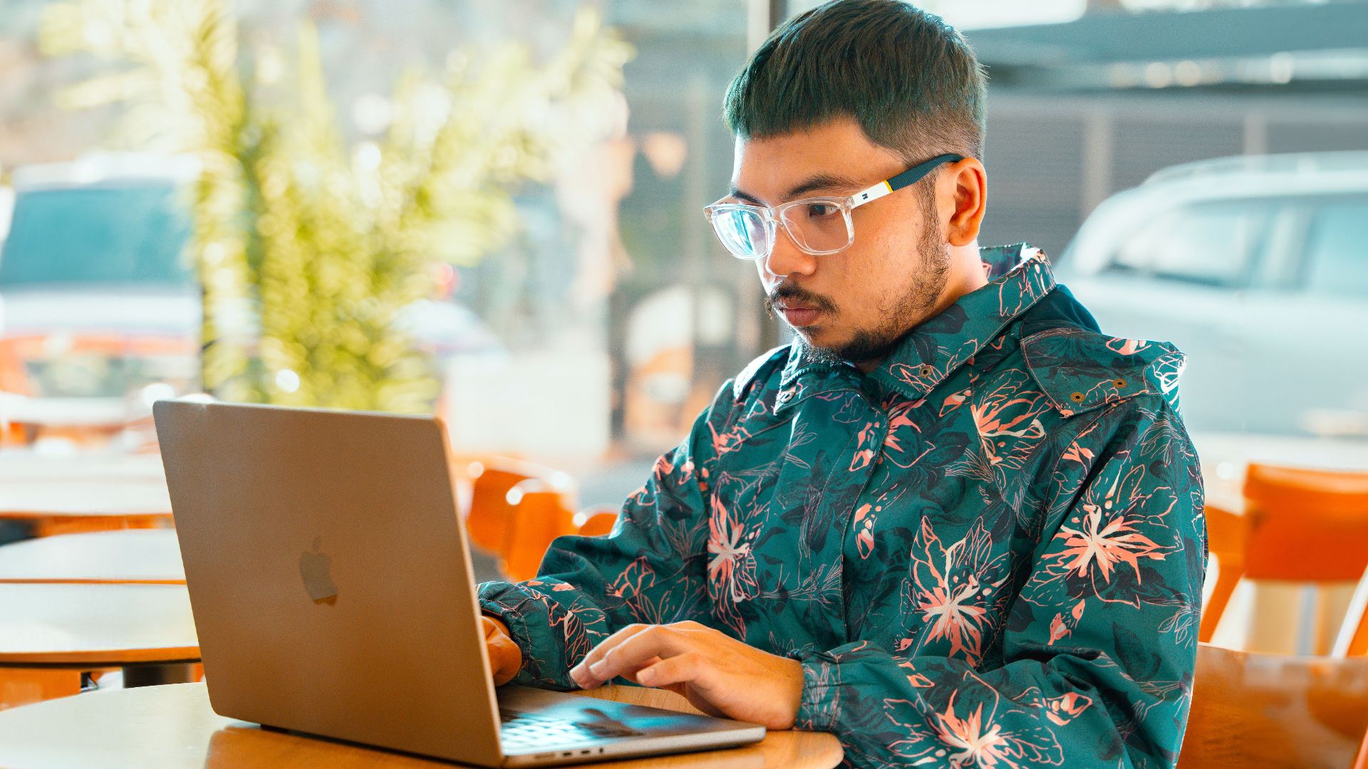 A young man focuses on his laptop in a bright Baguio cafe, wearing eyeglasses and a floral jacket.