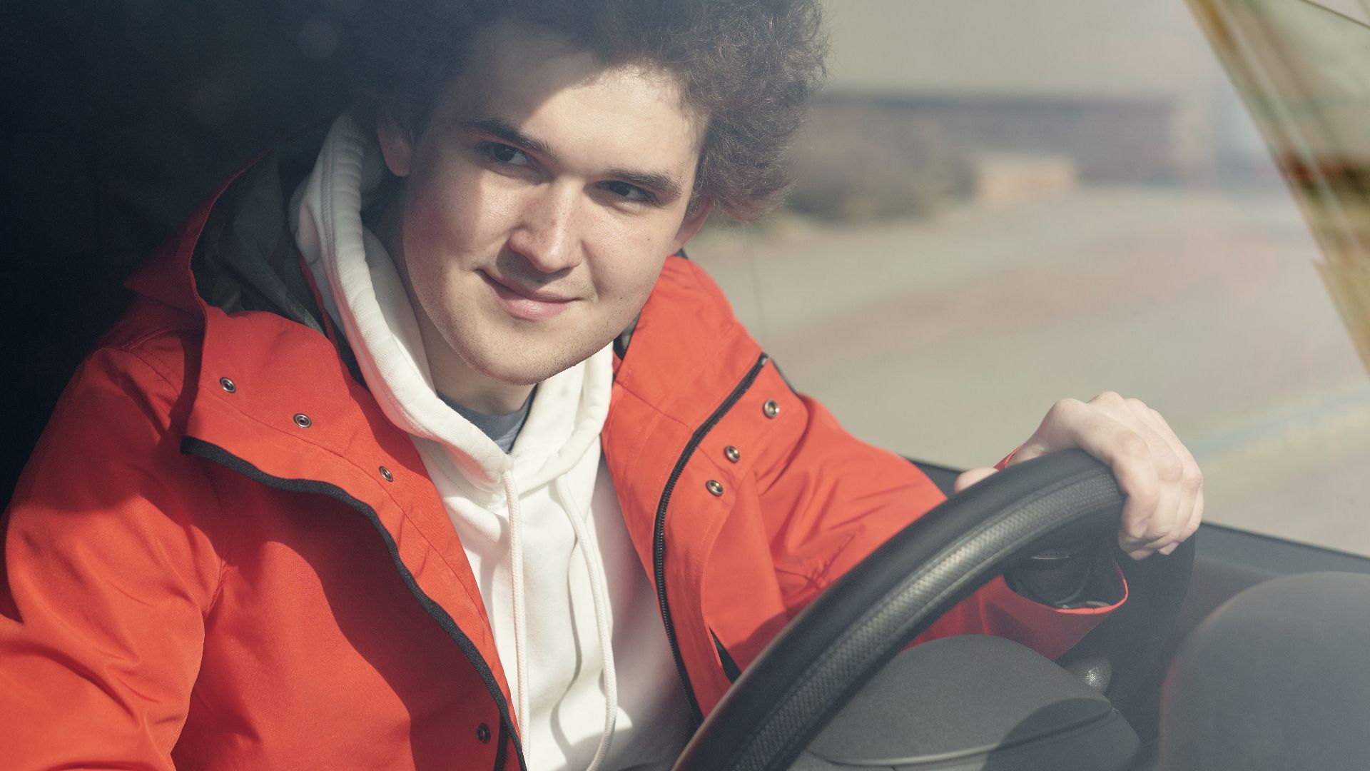 A young adult man smiling while sitting behind the steering wheel of a vehicle.