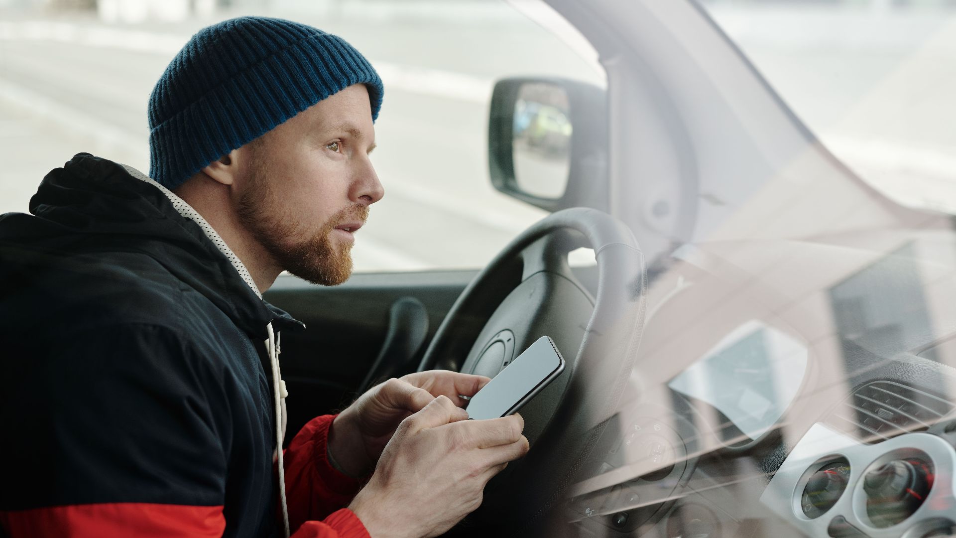Caucasian man in a knitted cap using a smartphone while sitting in a parked car.