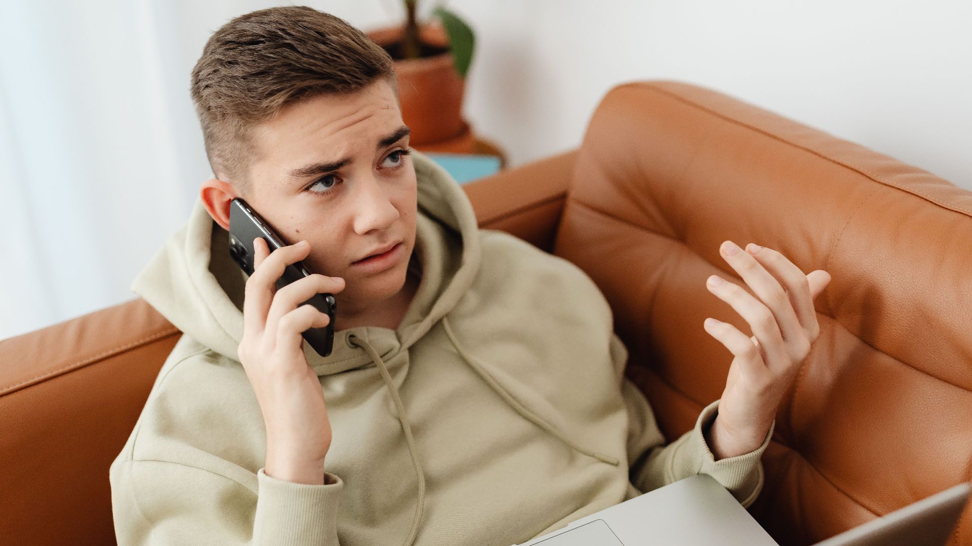 A young man talks on the phone while working on a laptop, looking concerned and frustrated.