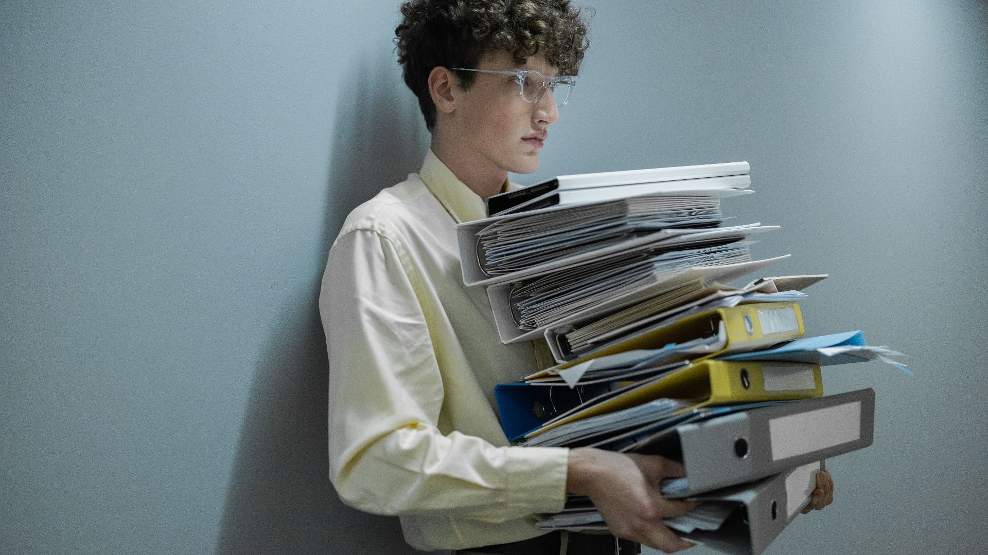 An overworked office worker carrying a large stack of files and folders in a modern setting.