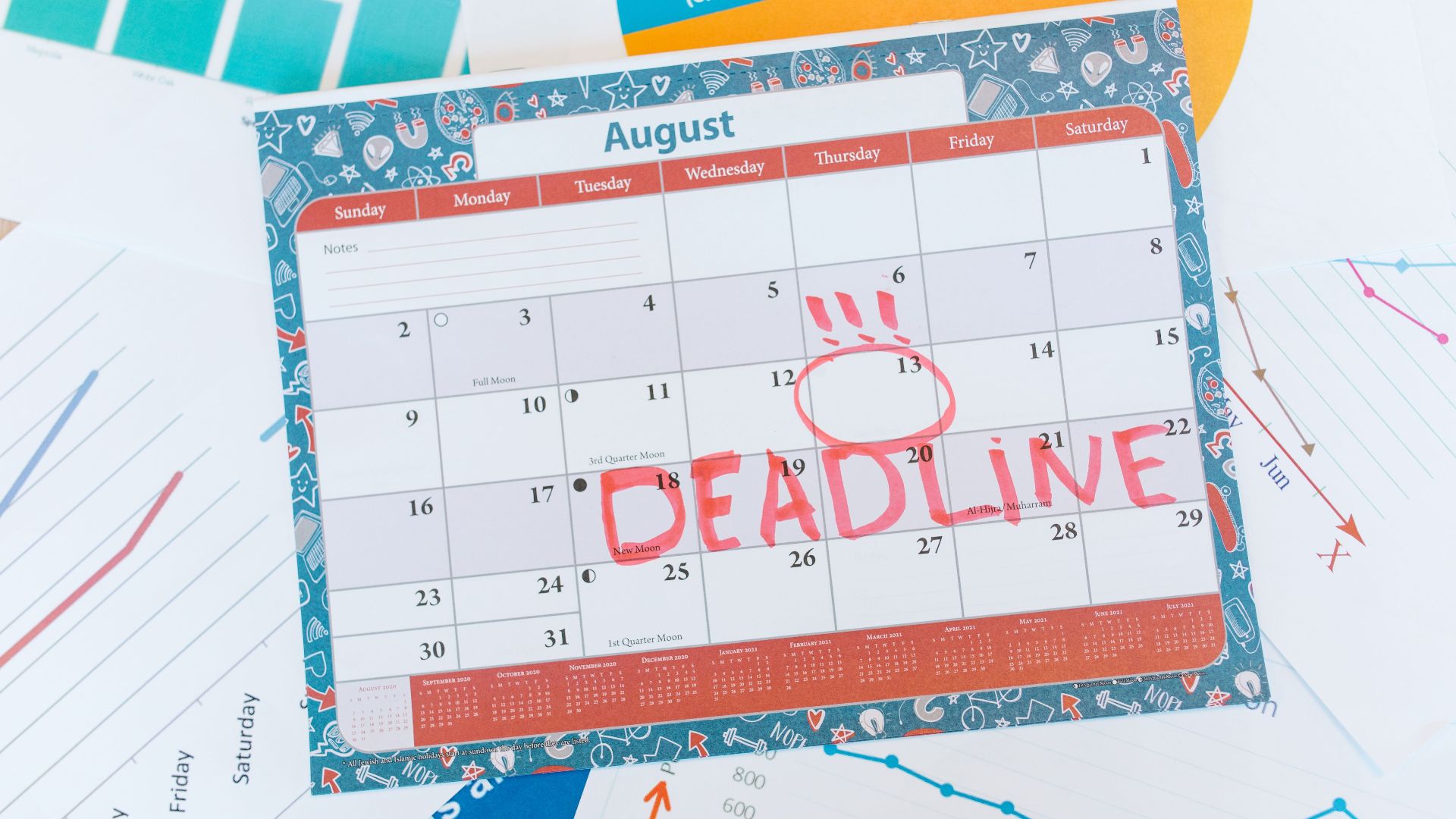 Vibrant August calendar on a desk with deadline marked in red, surrounded by graphs and charts.