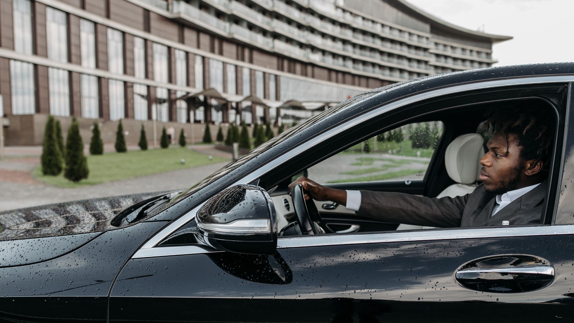 Black man driving a luxury vehicle in front of a modern building, exuding confidence.