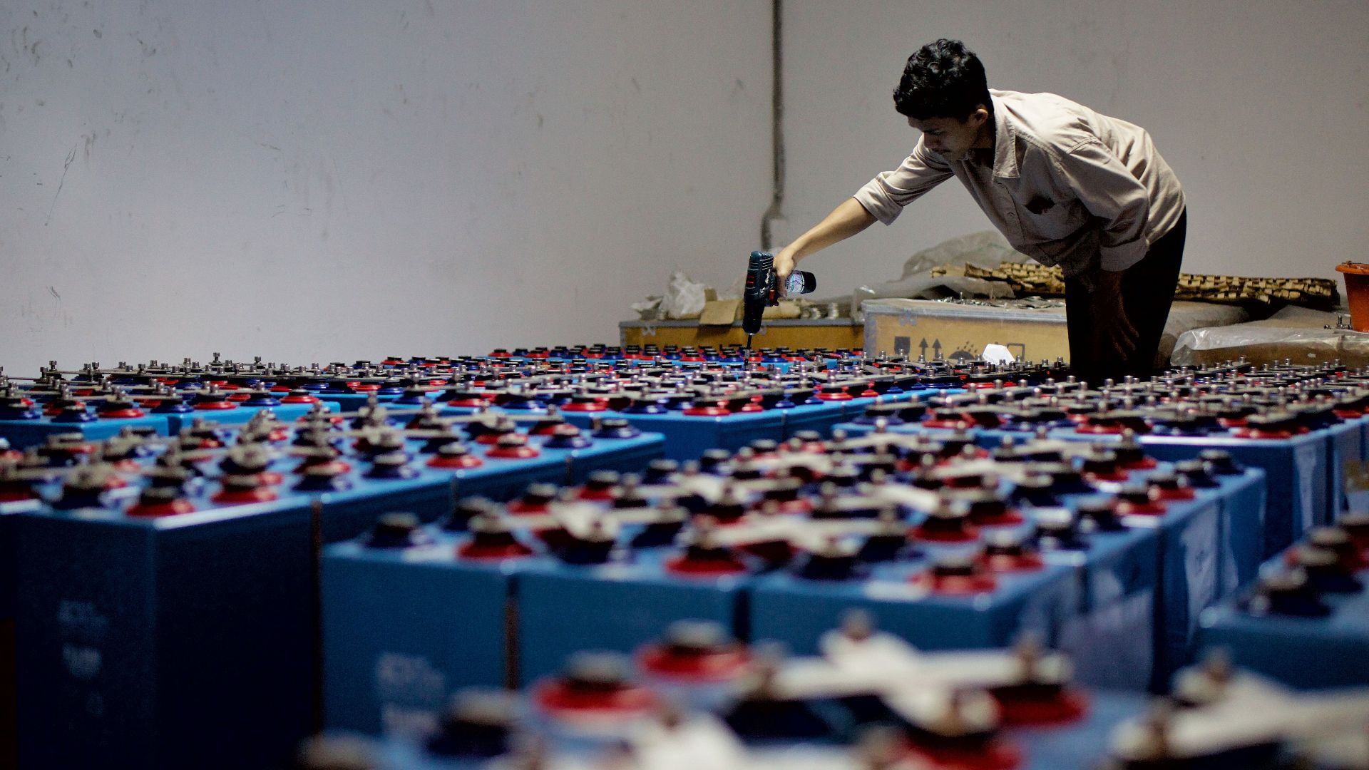 A worker checking many industrial batteries inside a facility. Indoor, industrial setting.