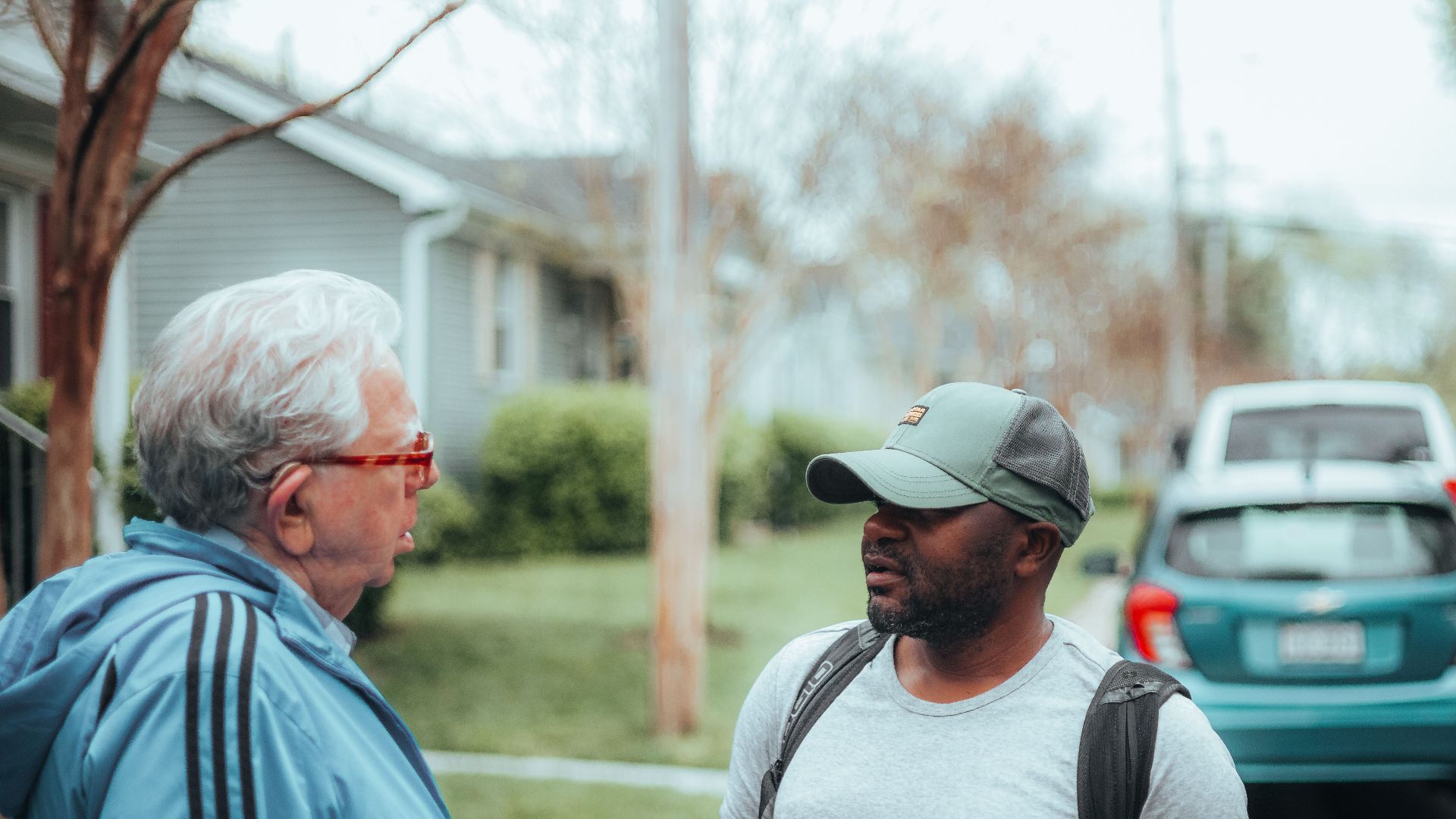 Two men engaged in conversation on a suburban sidewalk with parked cars.