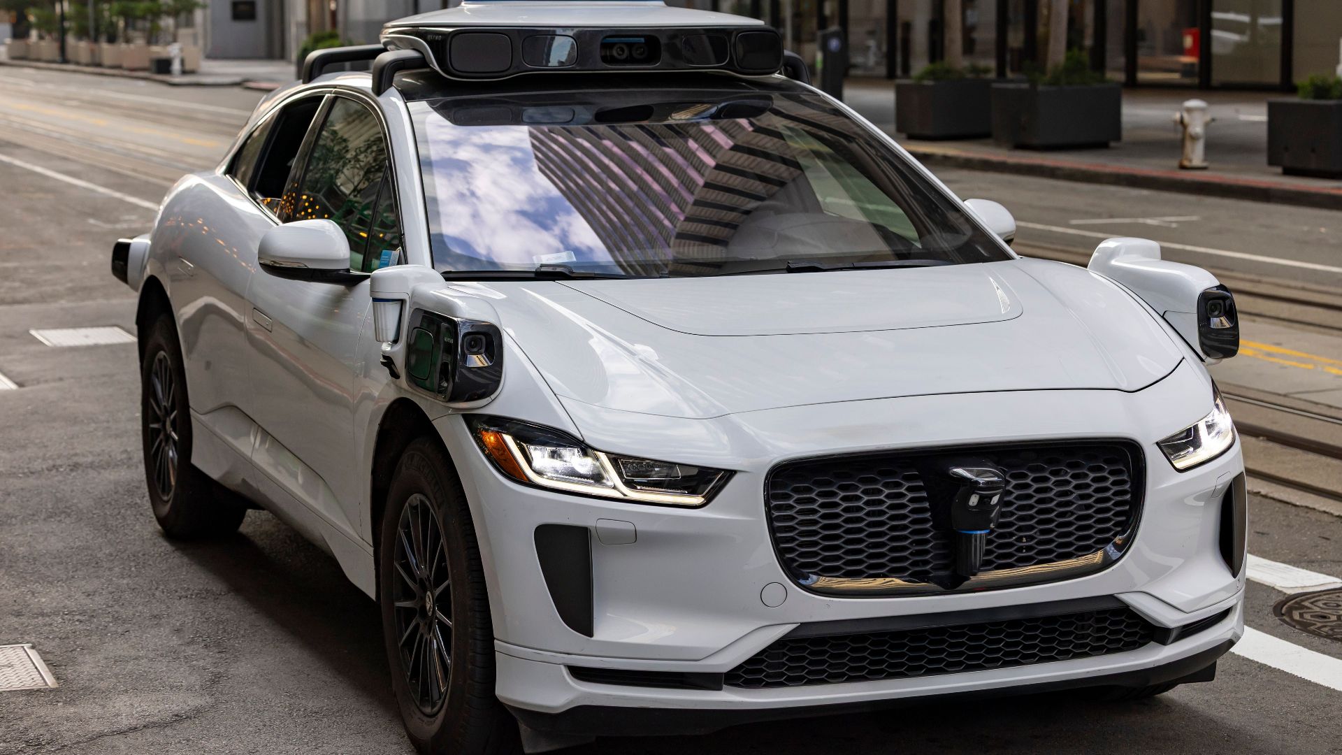 A white autonomous vehicle navigating a city street, reflecting urban architecture in daylight.