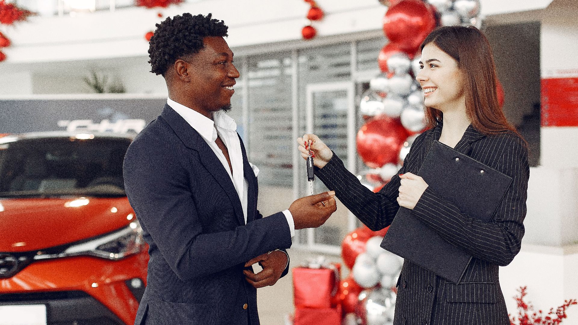 Side view of happy positive female dealer in formal classy wear passing car keys to  excited African American customer in trendy suit in car showroom