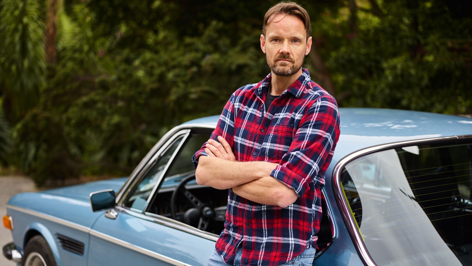 Adult bearded male in jeans and shirt standing with crossed arms leaned on blue retro auto near forest in daylight and looking at camera