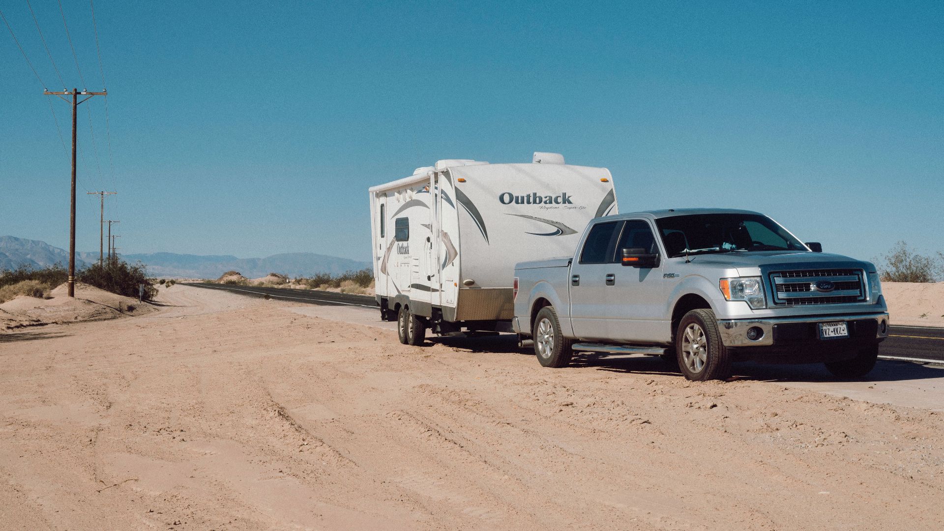 Silver truck towing an Outback trailer on a desert road under clear blue skies.