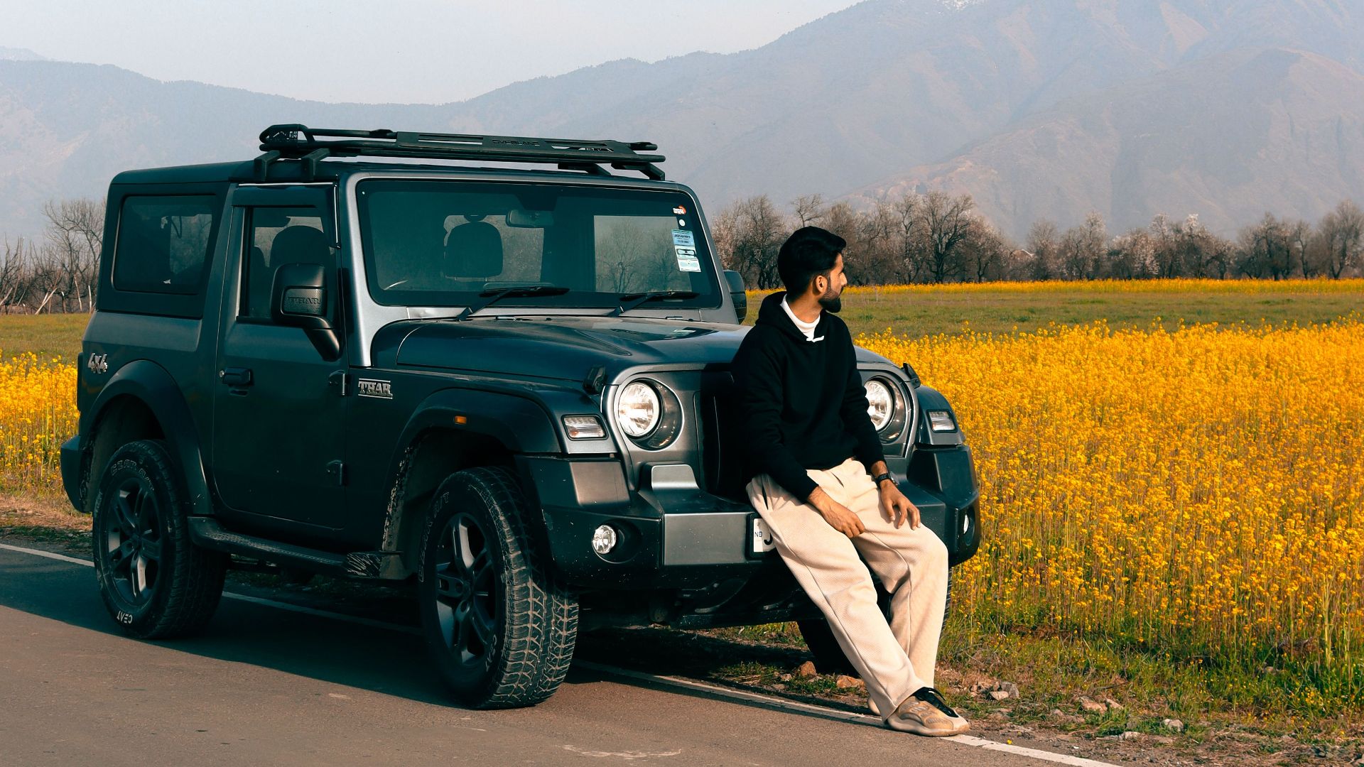 A man sits on a pick-up truck by a rural road with a backdrop of yellow flower fields and mountains.