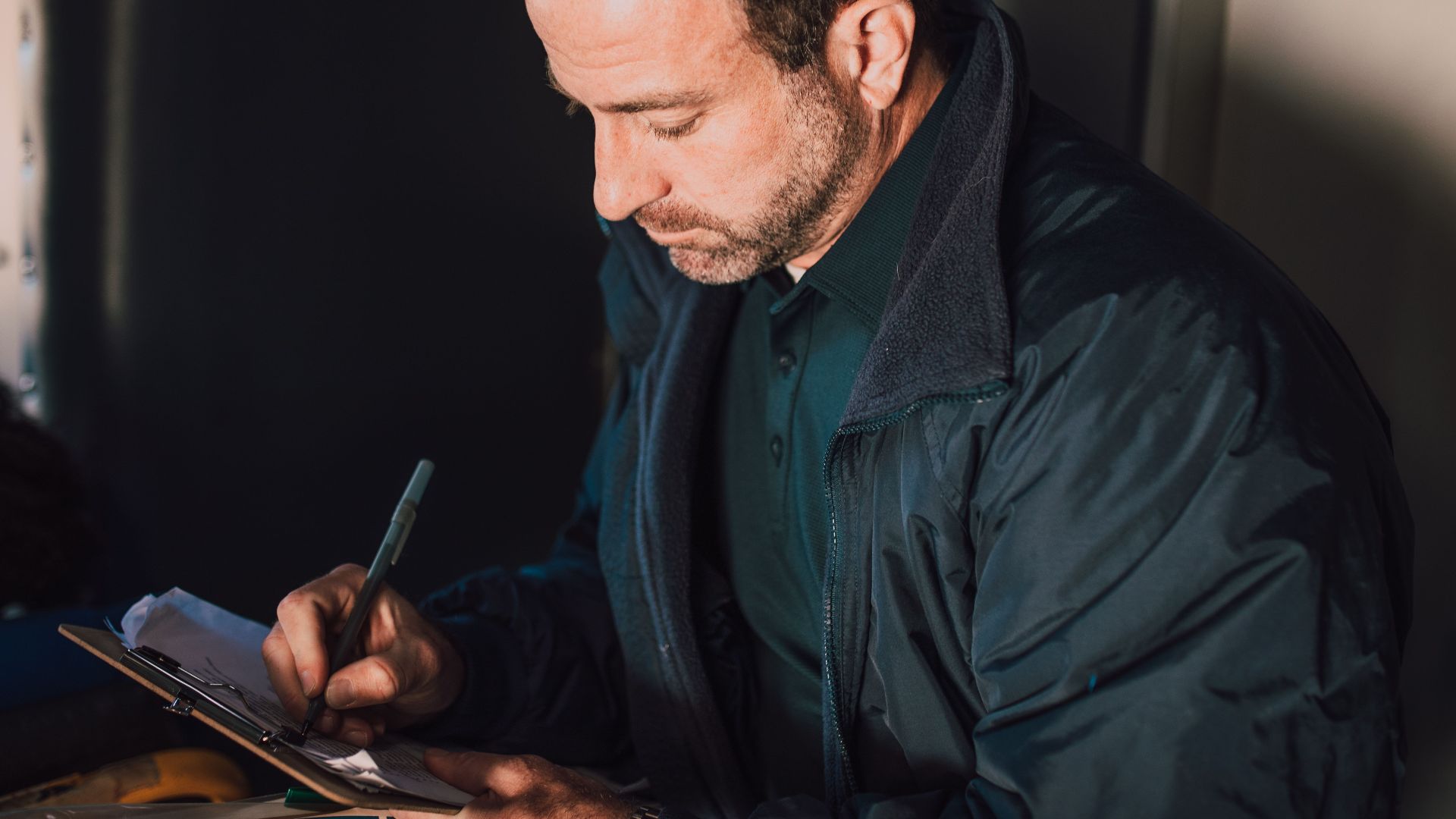 Adult male writing notes on a clipboard inside. Concentrated and thoughtful atmosphere.