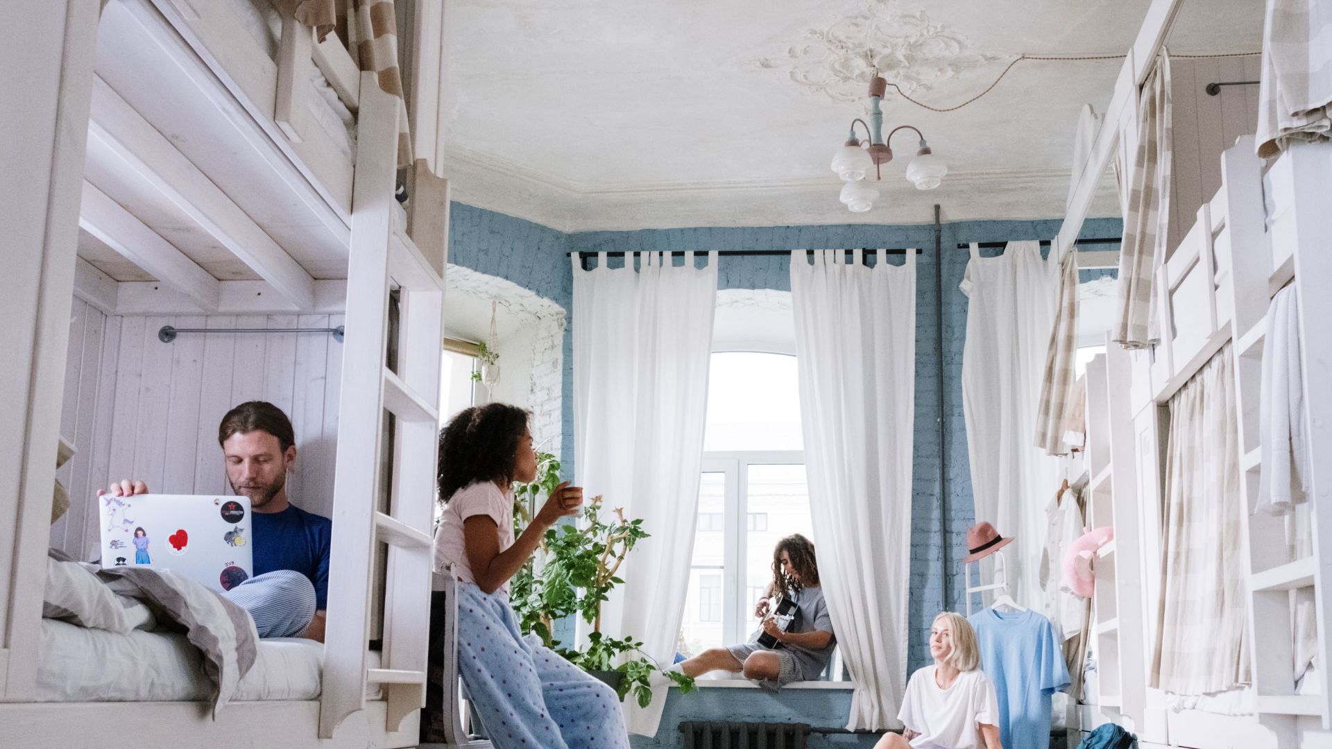 A cozy dorm room where friends relax, chat, play guitar, and use laptops together. A casual hangout environment.