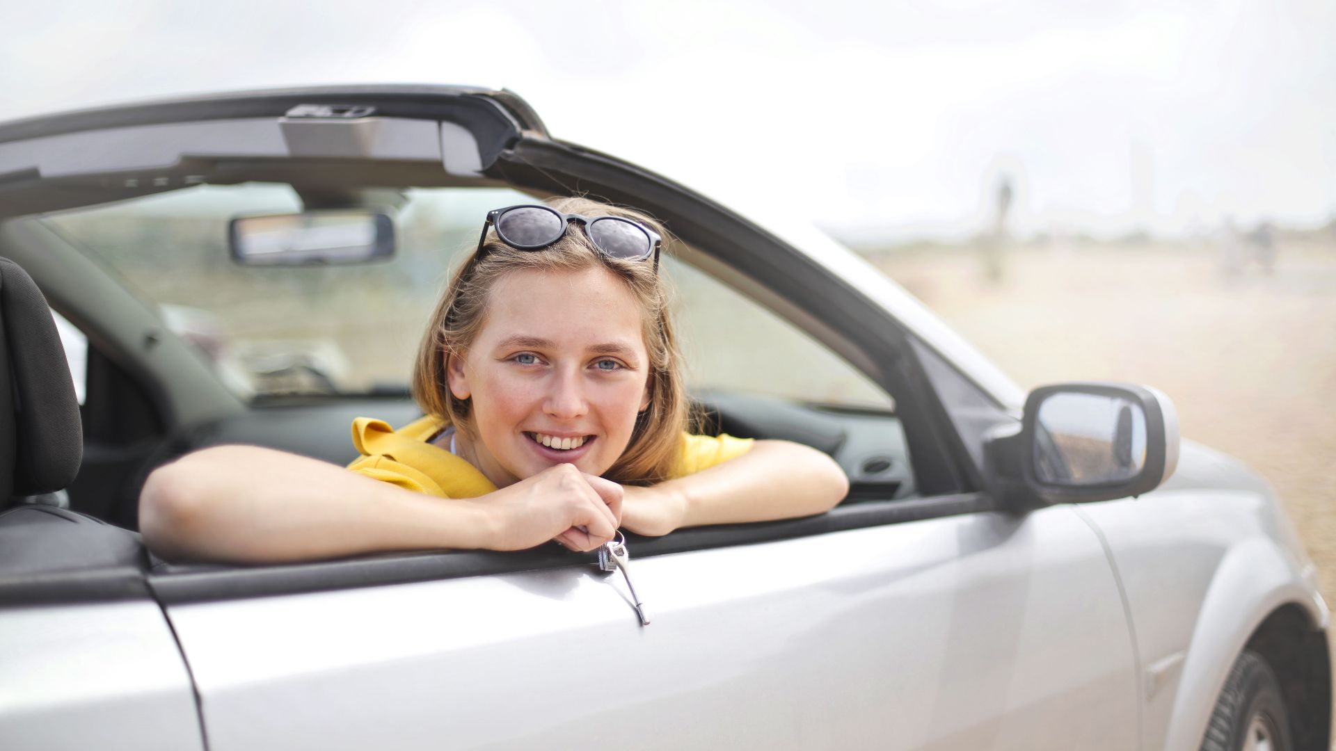 A young woman smiles joyfully while leaning out of a convertible car, enjoying a sunny day.