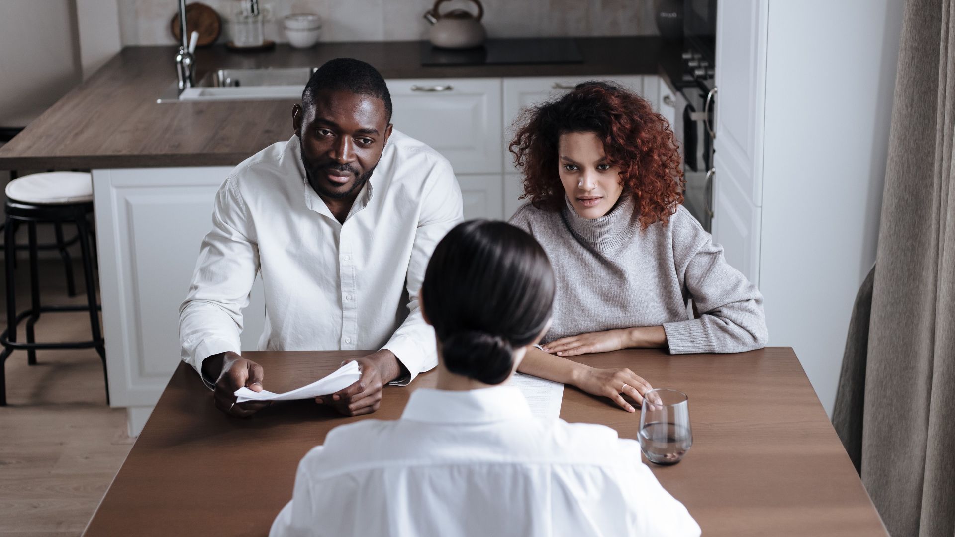 A couple attentively listens to an advisor during a meeting in a modern kitchen.