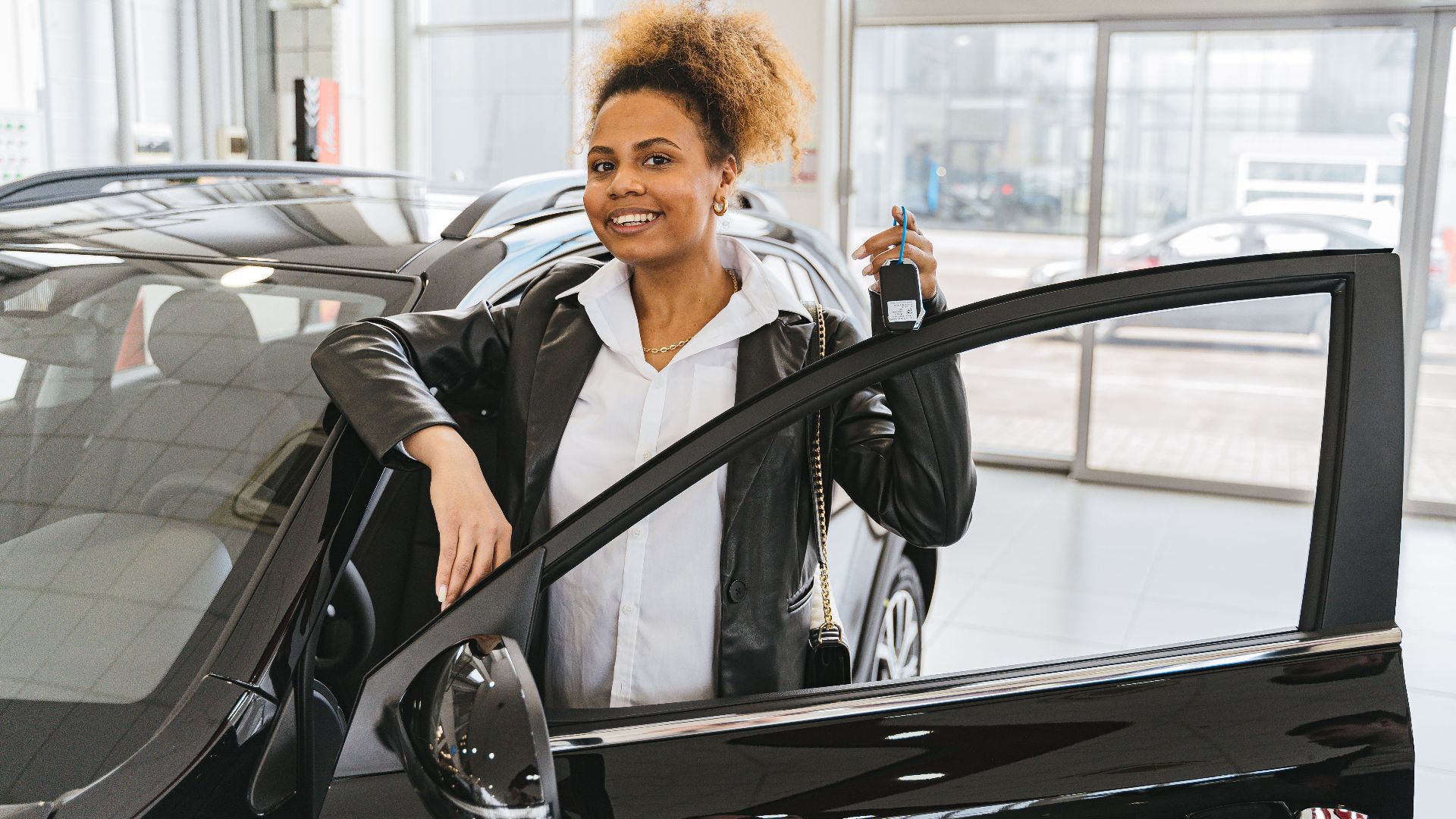 Smiling woman holding car key in dealership showroom next to new vehicle.
