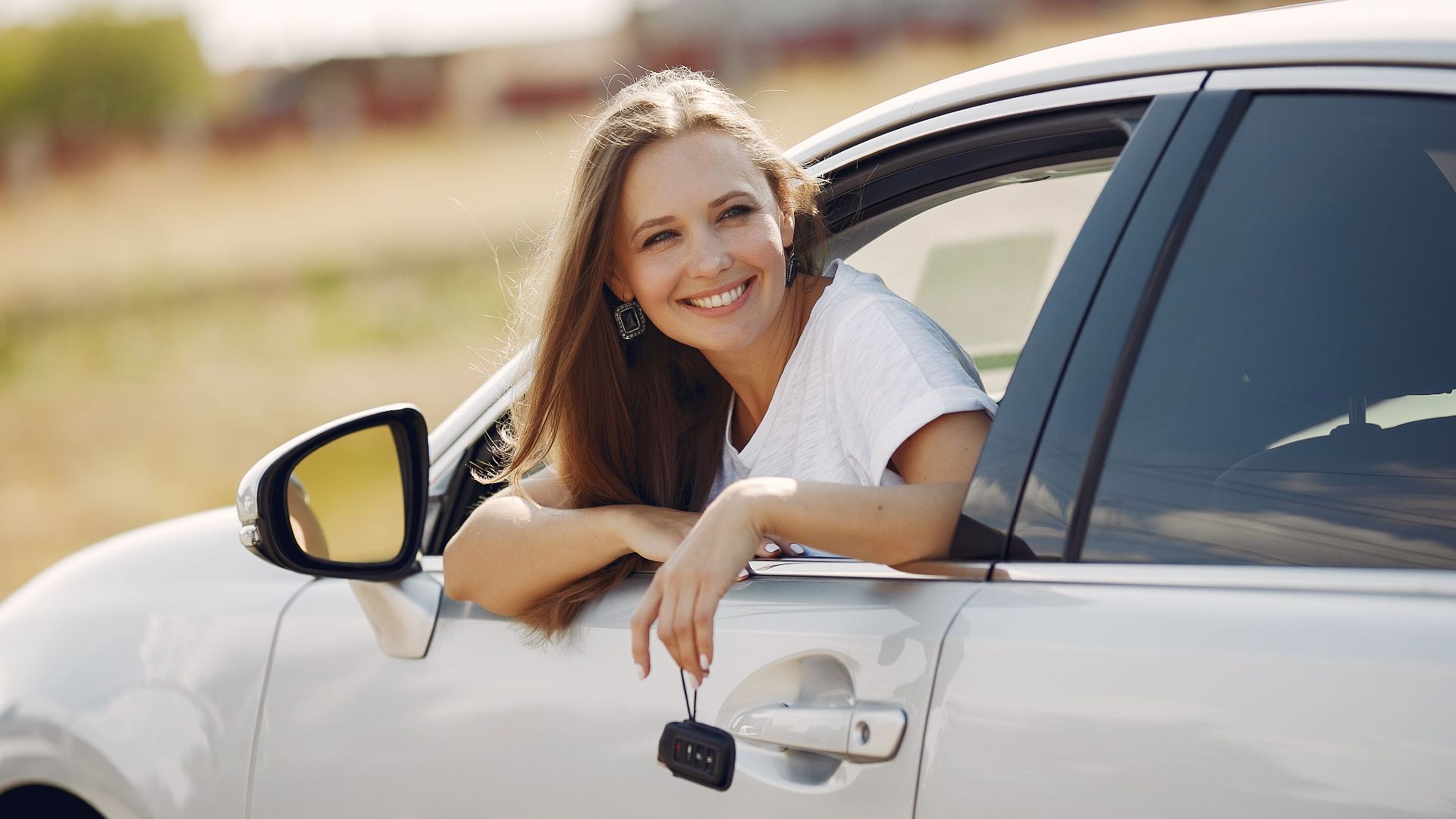 Happy female driver in white t shirt leaning out of side door window with car key in hand and looking away with smile in sunny summer day