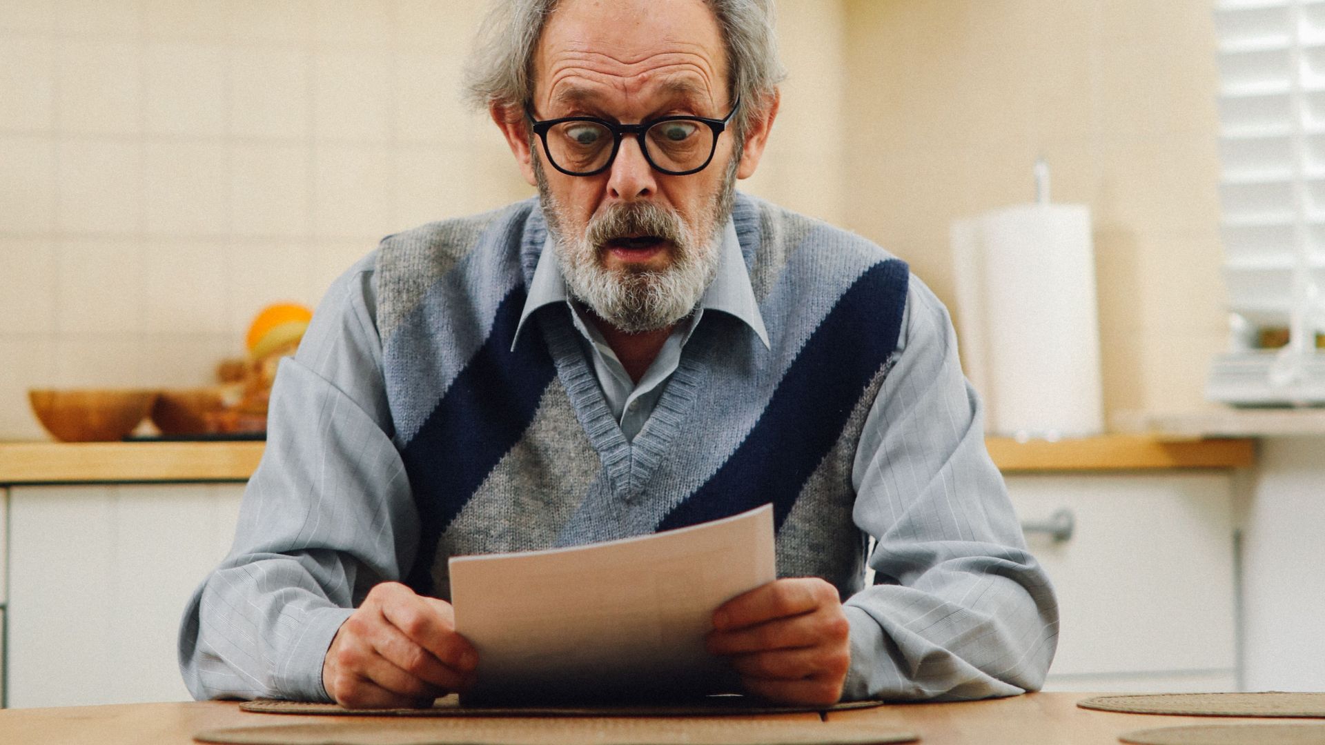 Senior man with eyeglasses reading a document at kitchen table, reflecting surprise.