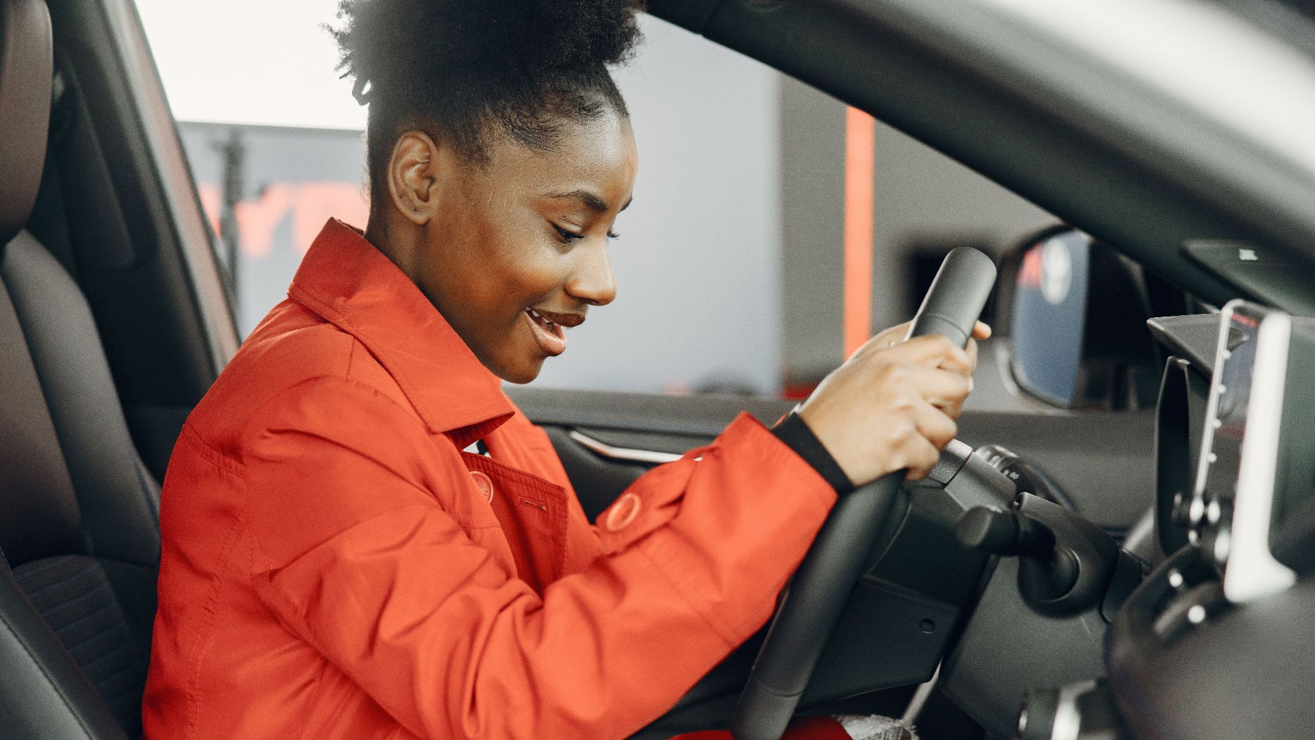 Young woman in an orange jacket enthusiastically trying out a car's steering wheel.