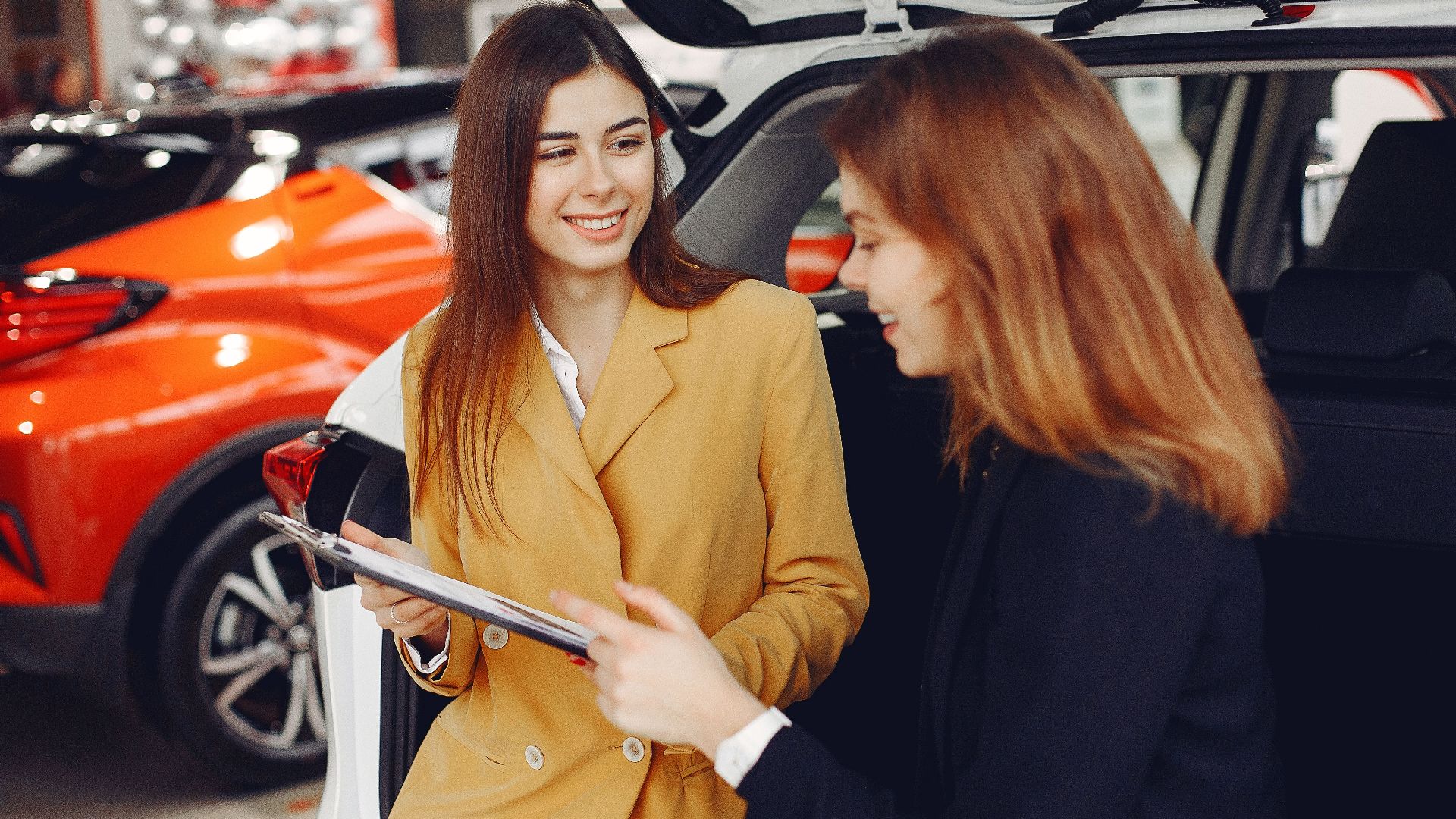 Positive smiling agent in trendy formal light brown suit discussing with happy excited female customer details of contract while leaning on car in showroom