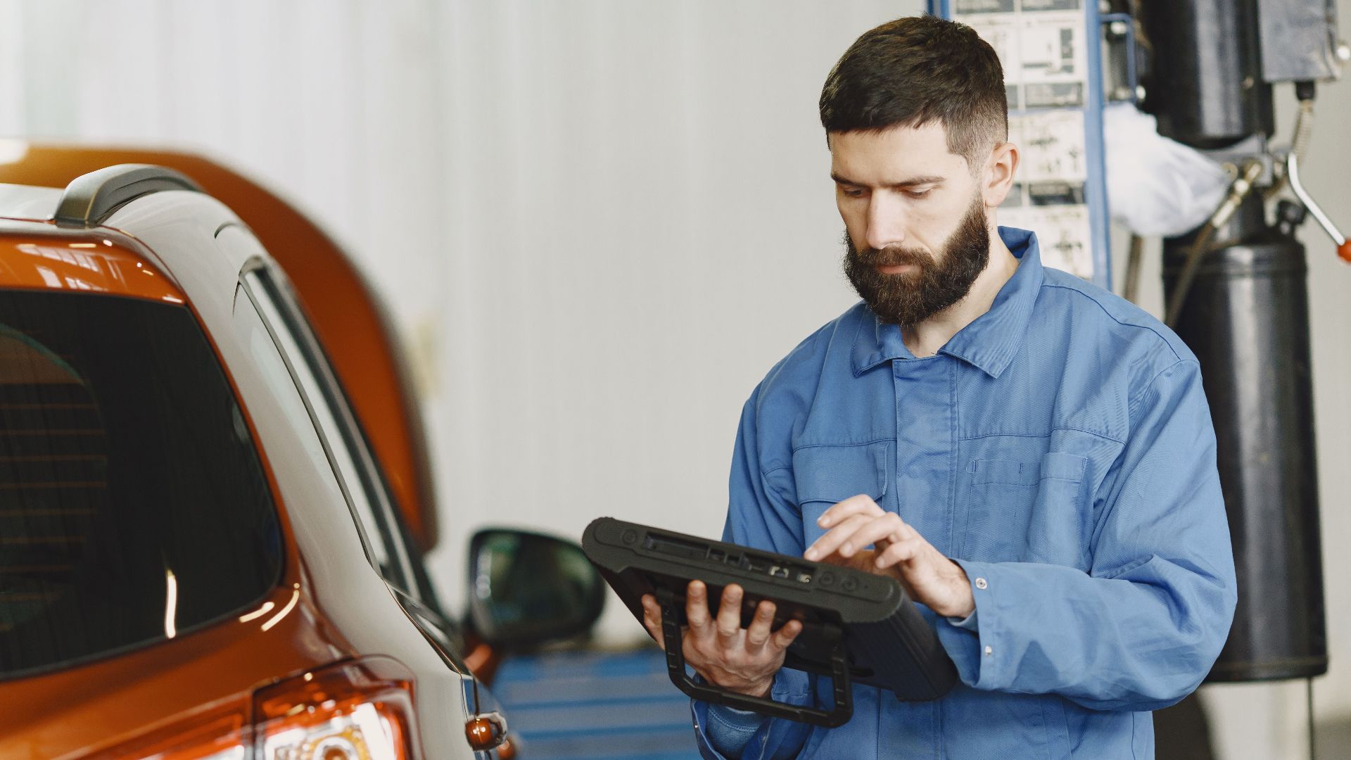 A mechanic in coveralls using a diagnostic tool at an auto repair shop beside a car.