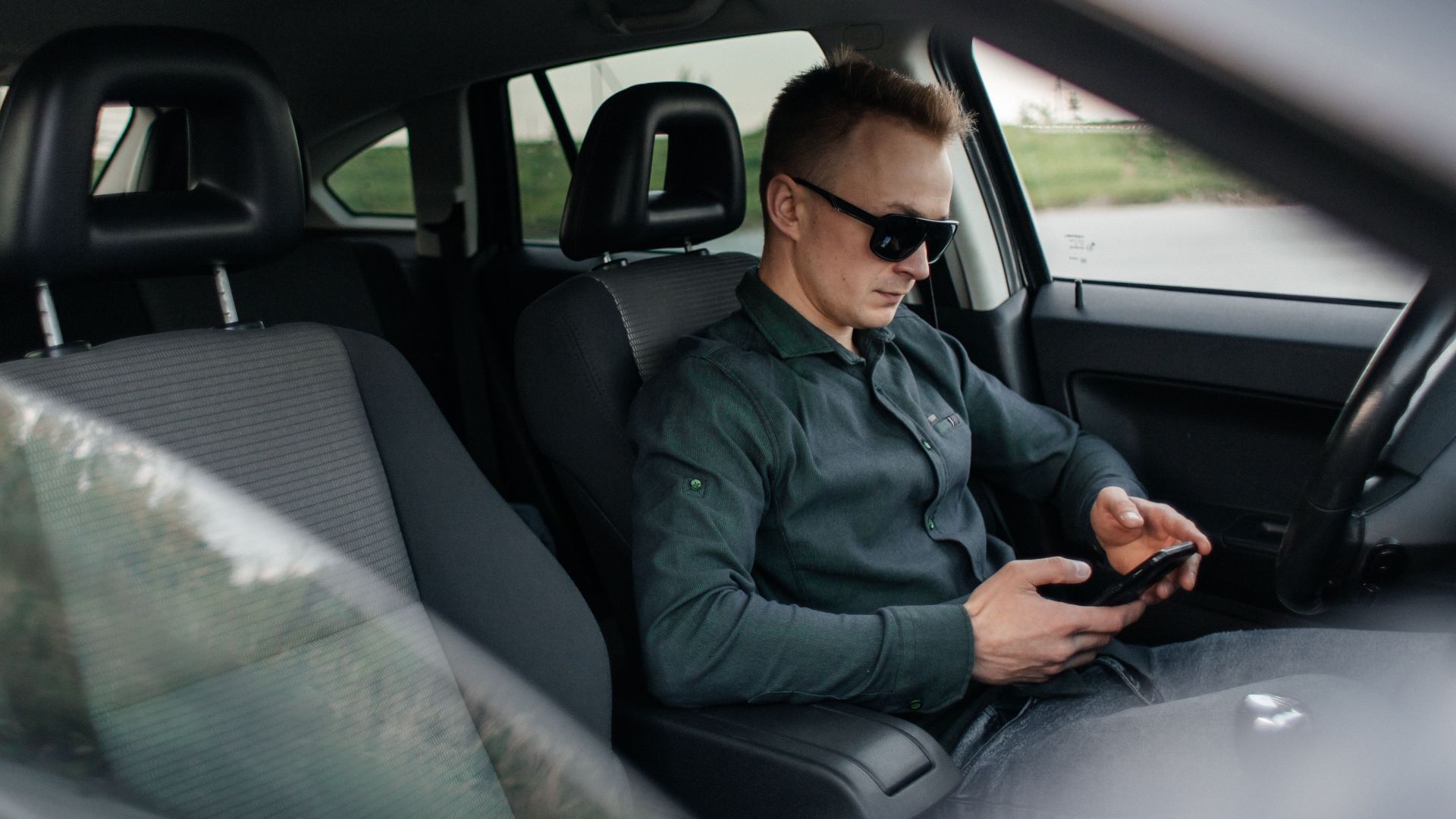 Young man using smartphone inside a parked car, wearing sunglasses.