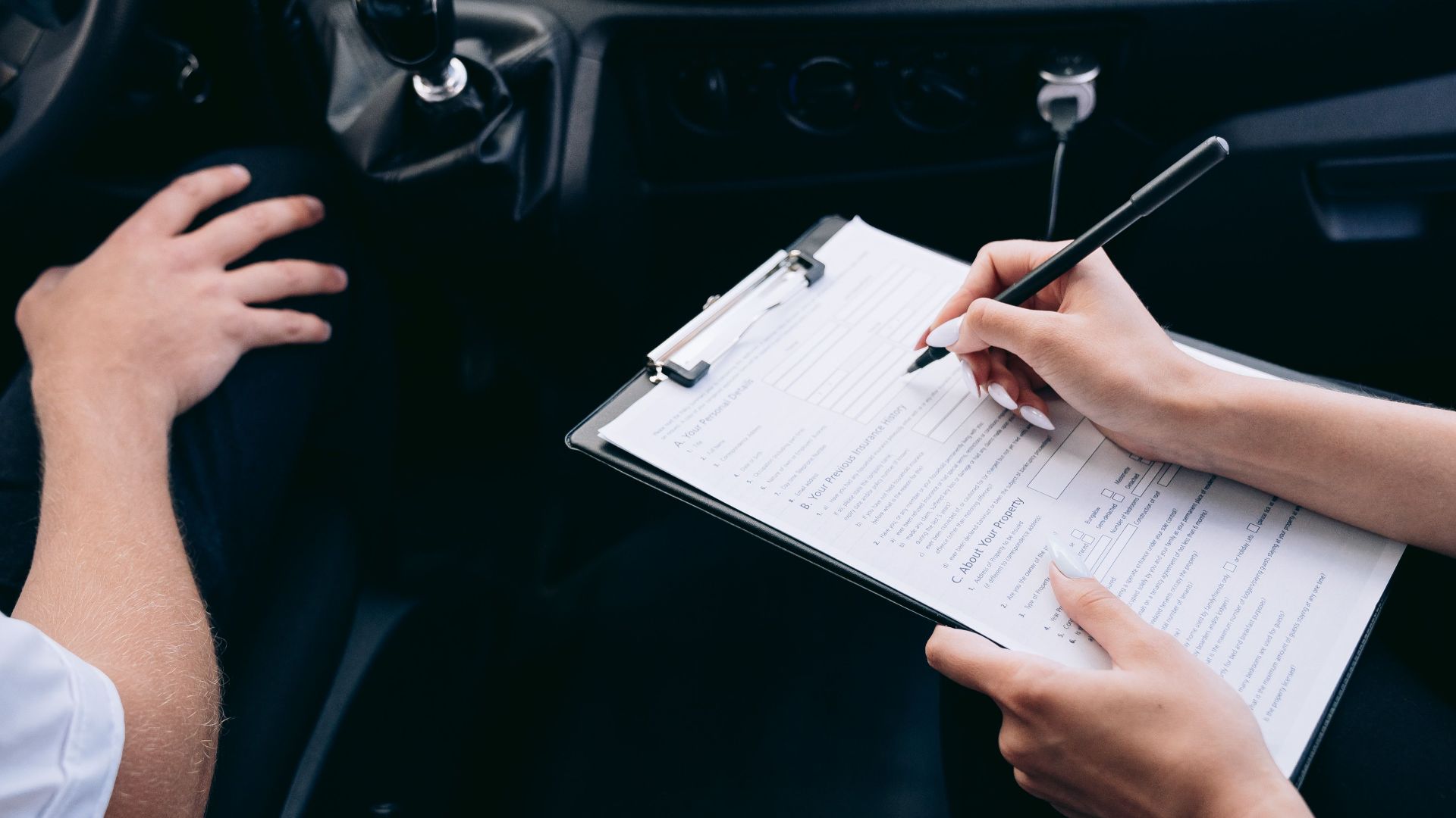 Close-up of a person writing on a clipboard inside a car, showing hands and a gear shift.