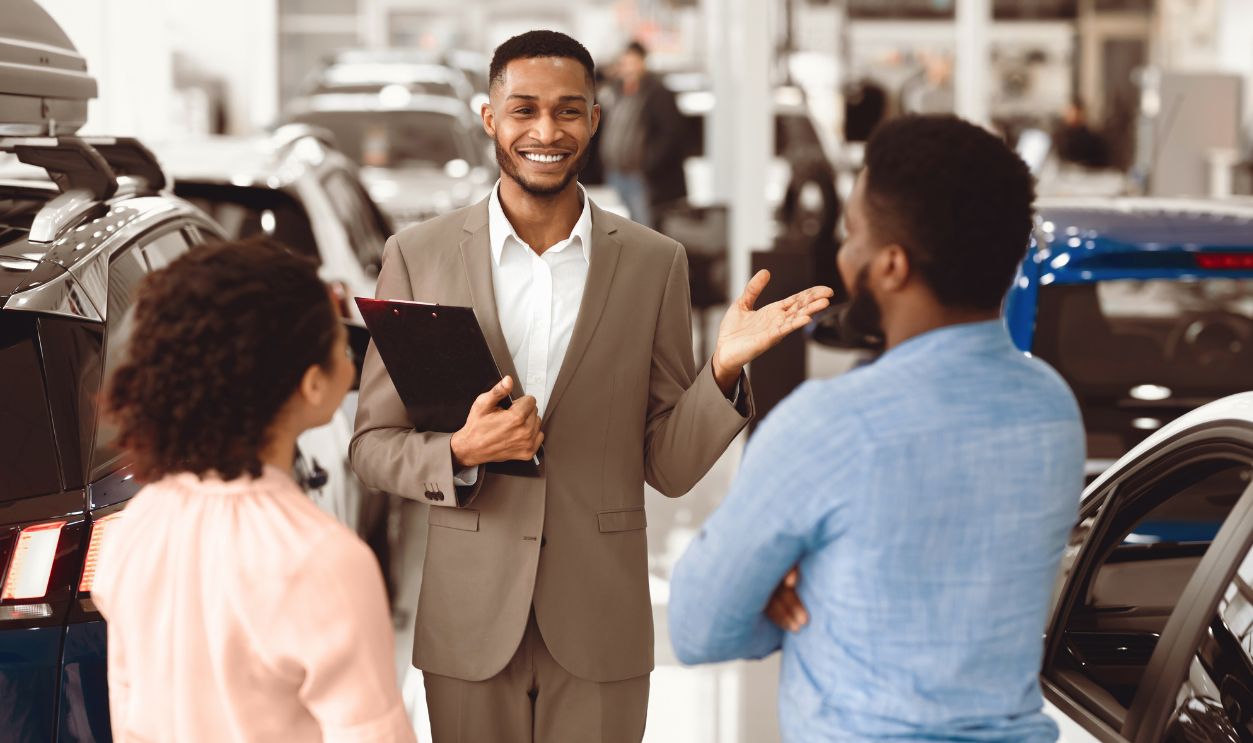 Professional Car Seller Talking With African American Couple Clients Selling Auto Working In Dealership Center. Selective Focus