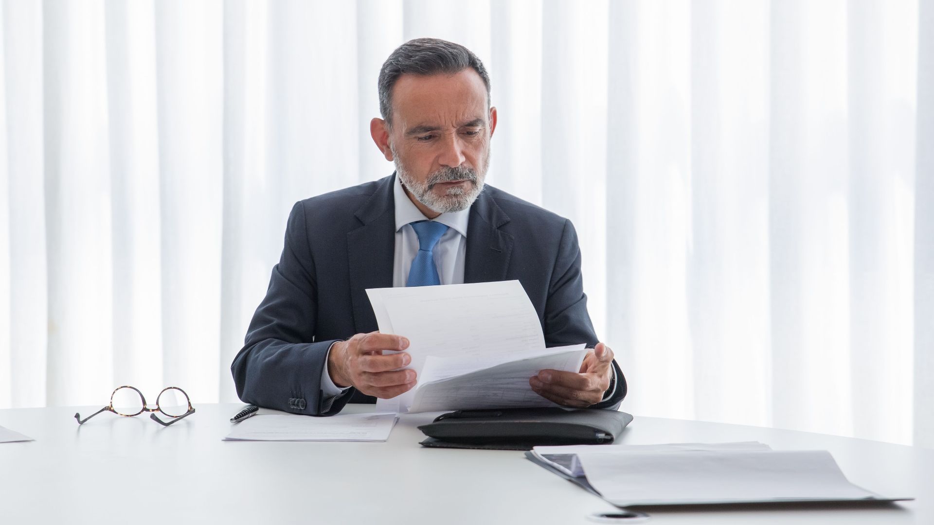 Elderly businessman focusing on reading documents at a table in a bright office setting.