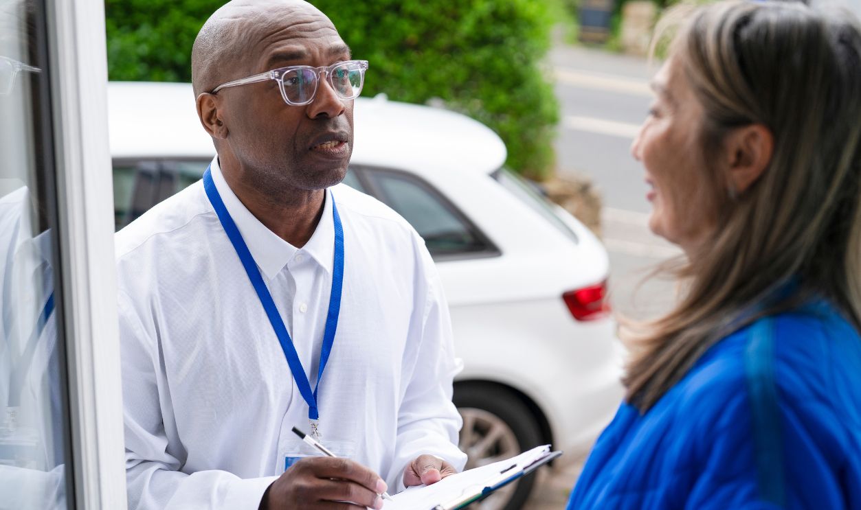 A man stands on the porch talking to a woman. He holds a clipboard and seems to be asking questions. There are cars parked nearby and trees in the background.