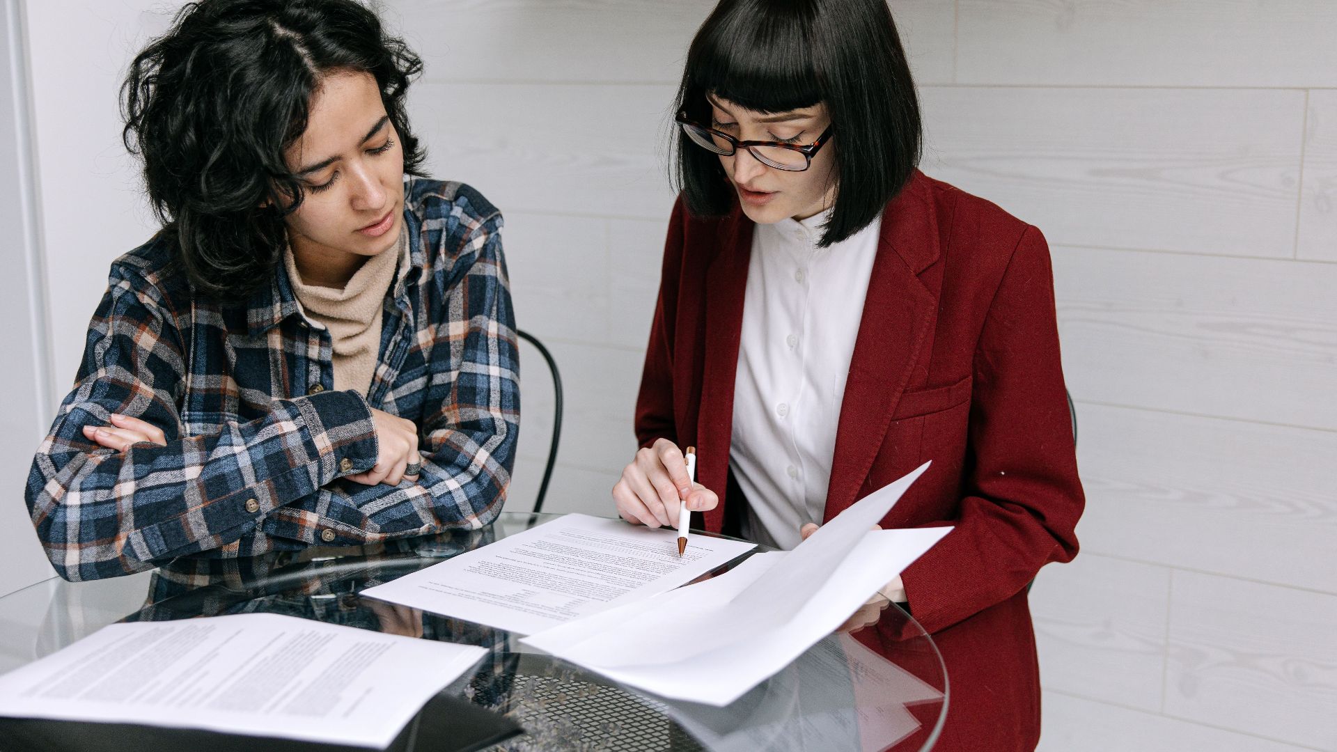 Two women collaborating at a glass table, reviewing business documents in a modern office setting.