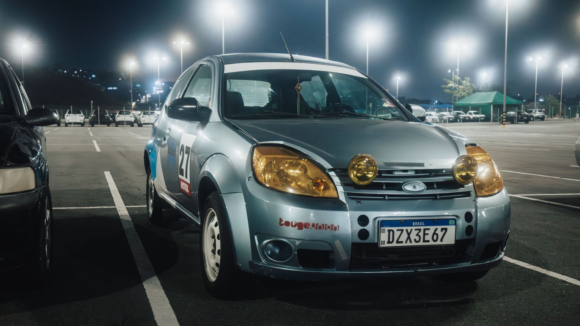Silver car with yellow headlights in a lit parking lot at night.
