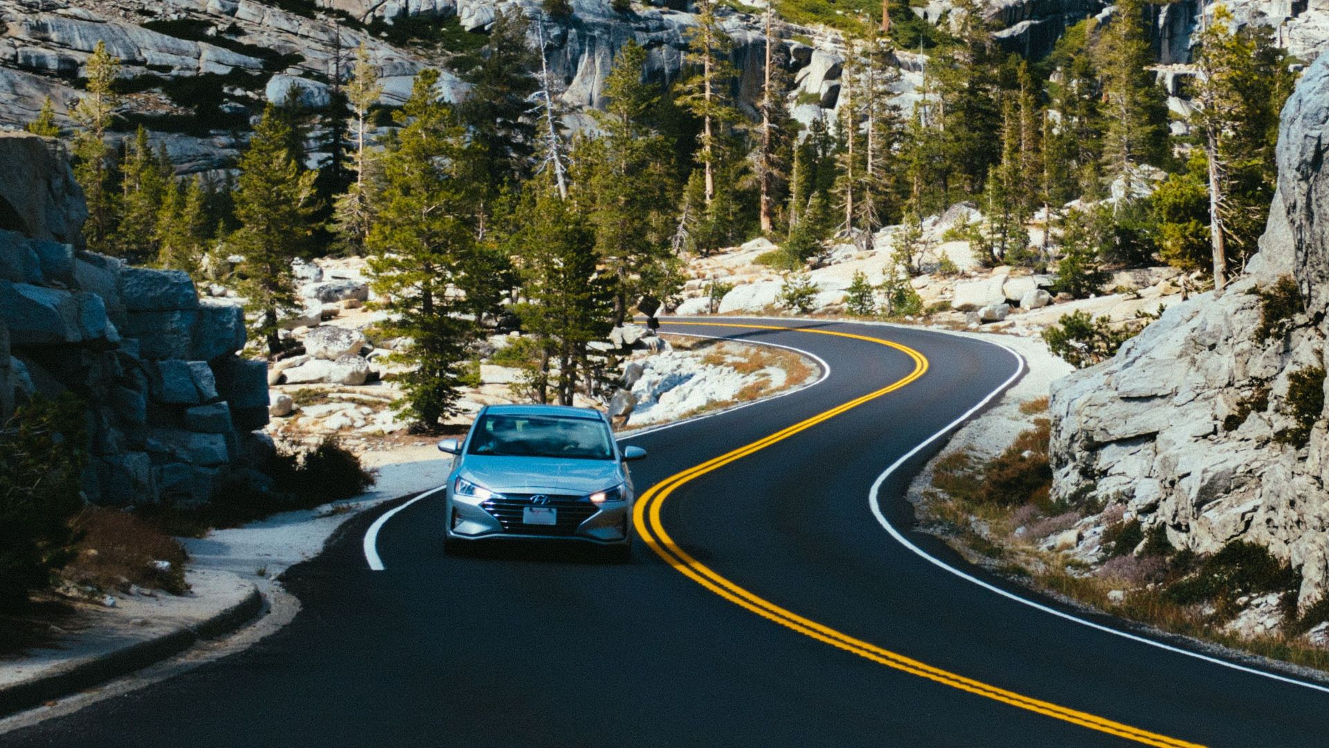 A car drives through a winding road in a mountainous landscape, surrounded by trees and rocks under a clear summer sky.