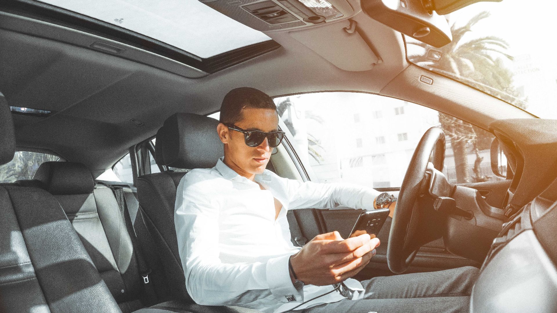 A man wearing sunglasses texts on his phone while driving a car in Morocco.