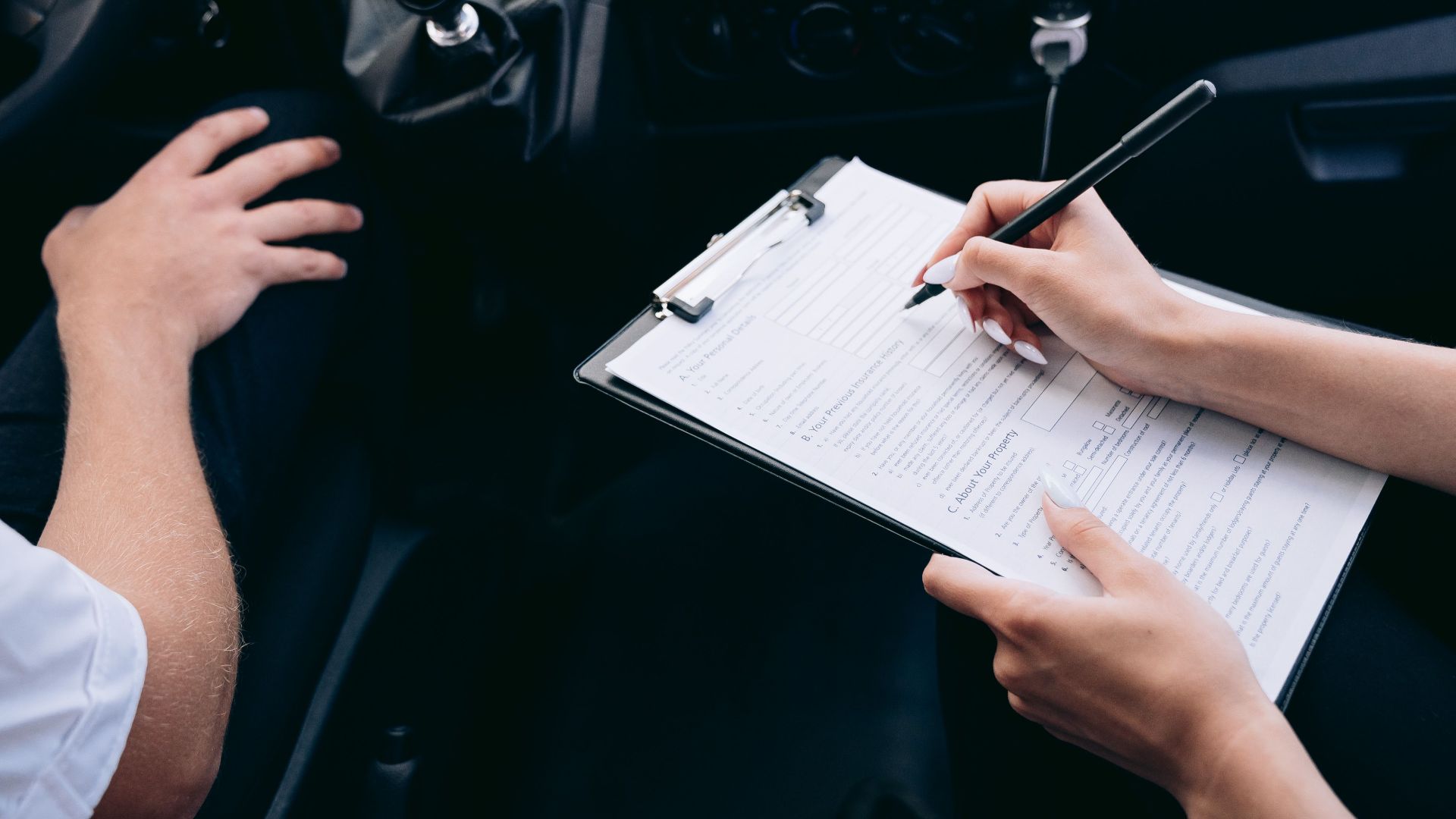Close-up of a person writing on a clipboard inside a car, showing hands and a gear shift.