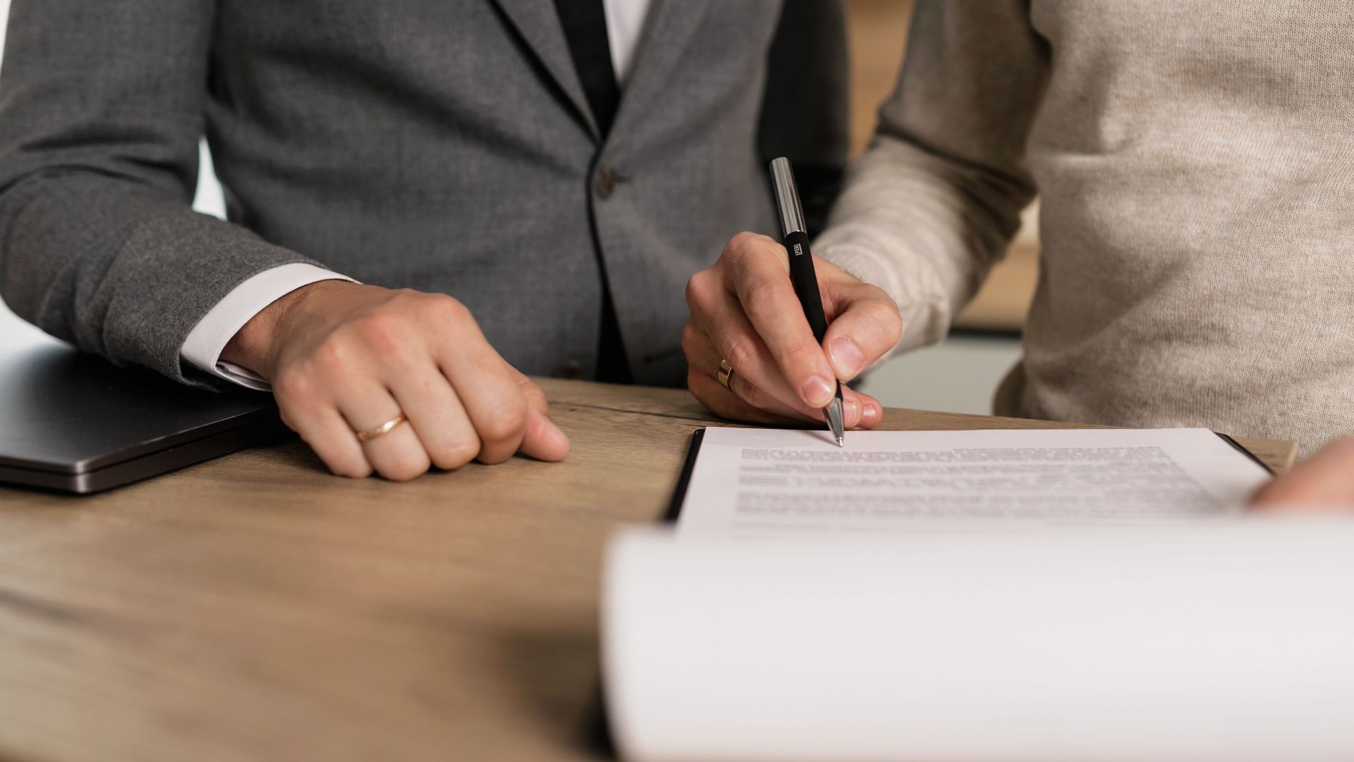 Close-up of businessmen signing documents at a wooden table in an office.