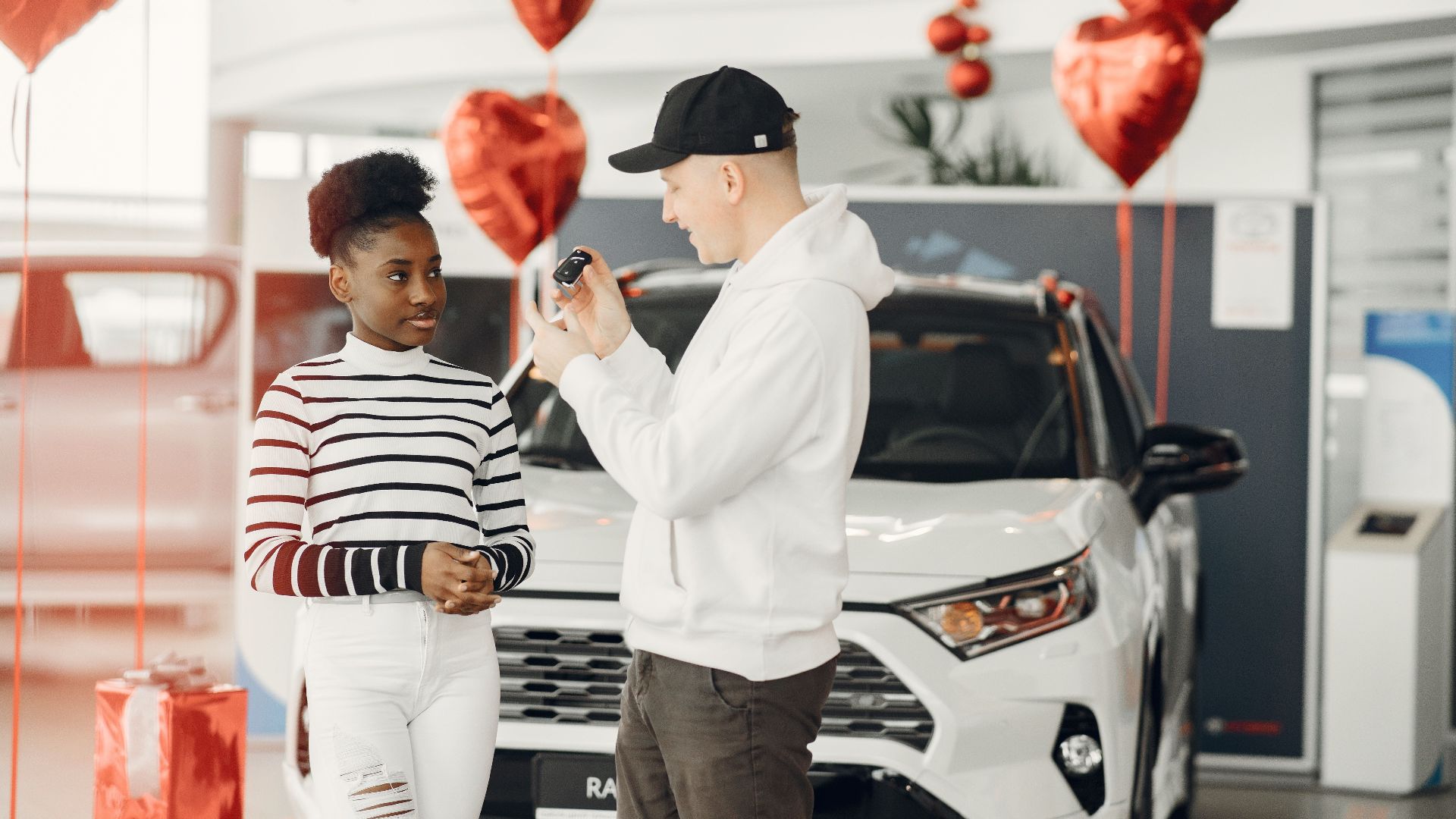 A couple exchanging car keys inside a showroom decorated with heart-shaped balloons, celebrating a new car purchase.