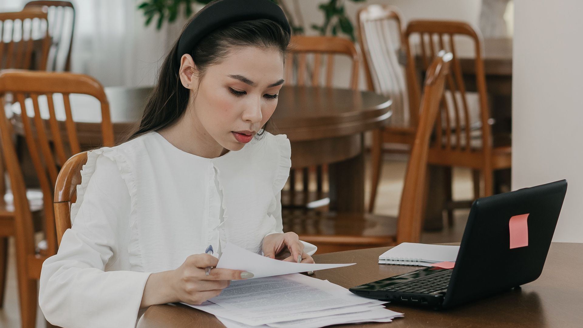 Focused woman studying with papers and laptop in a quiet library setting, surrounded by wooden tables and chairs.