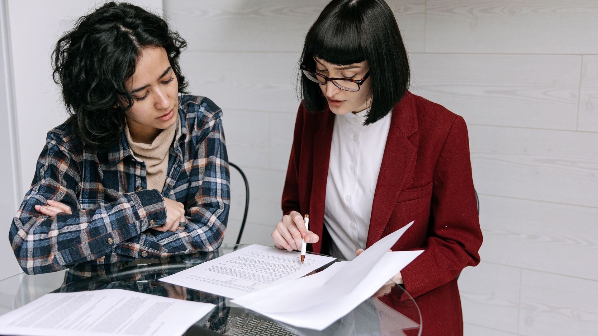 Two women collaborating at a glass table, reviewing business documents in a modern office setting.