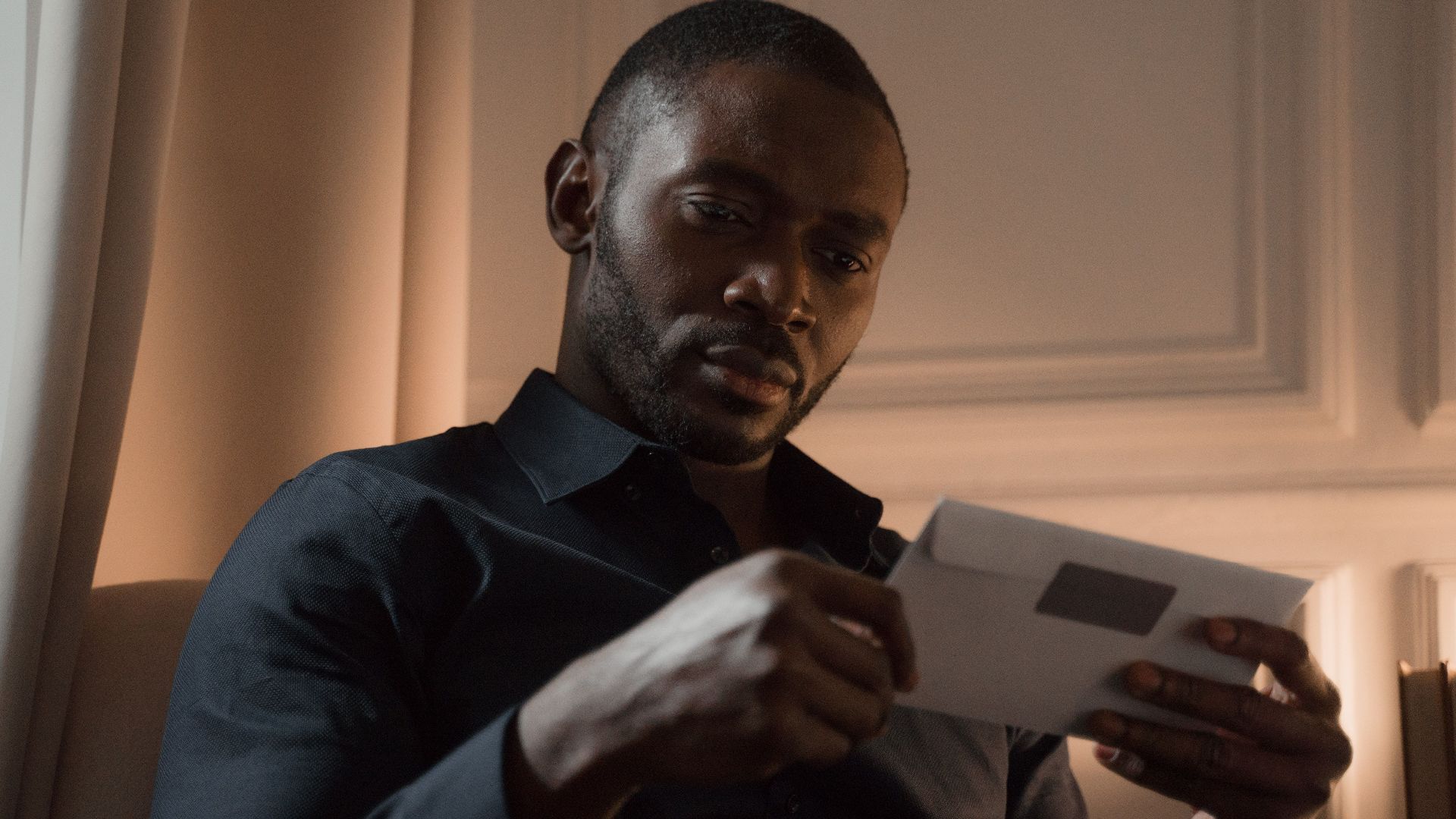 A man with a mustache reads a letter in a warmly lit room, creating a contemplative mood.