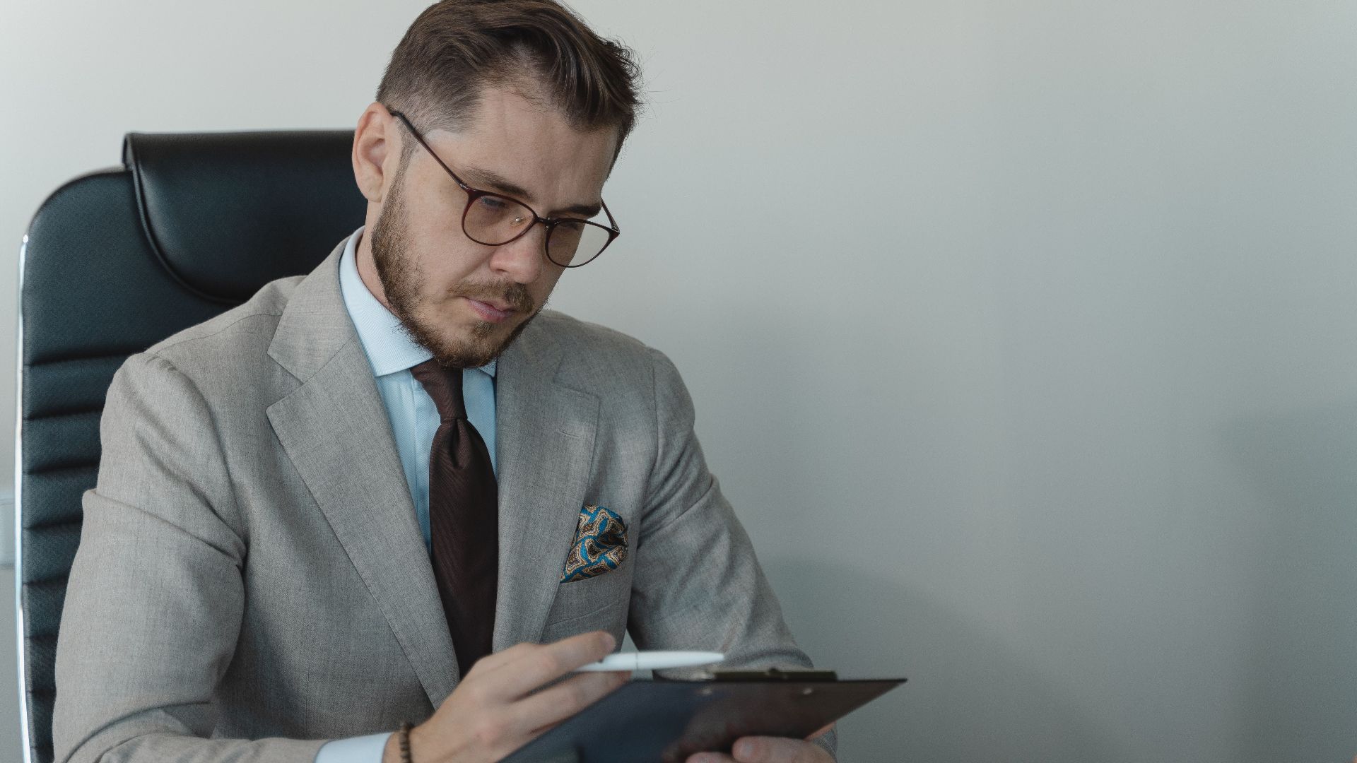 Businessman in a gray suit with glasses reviewing documents on a clipboard in an office.