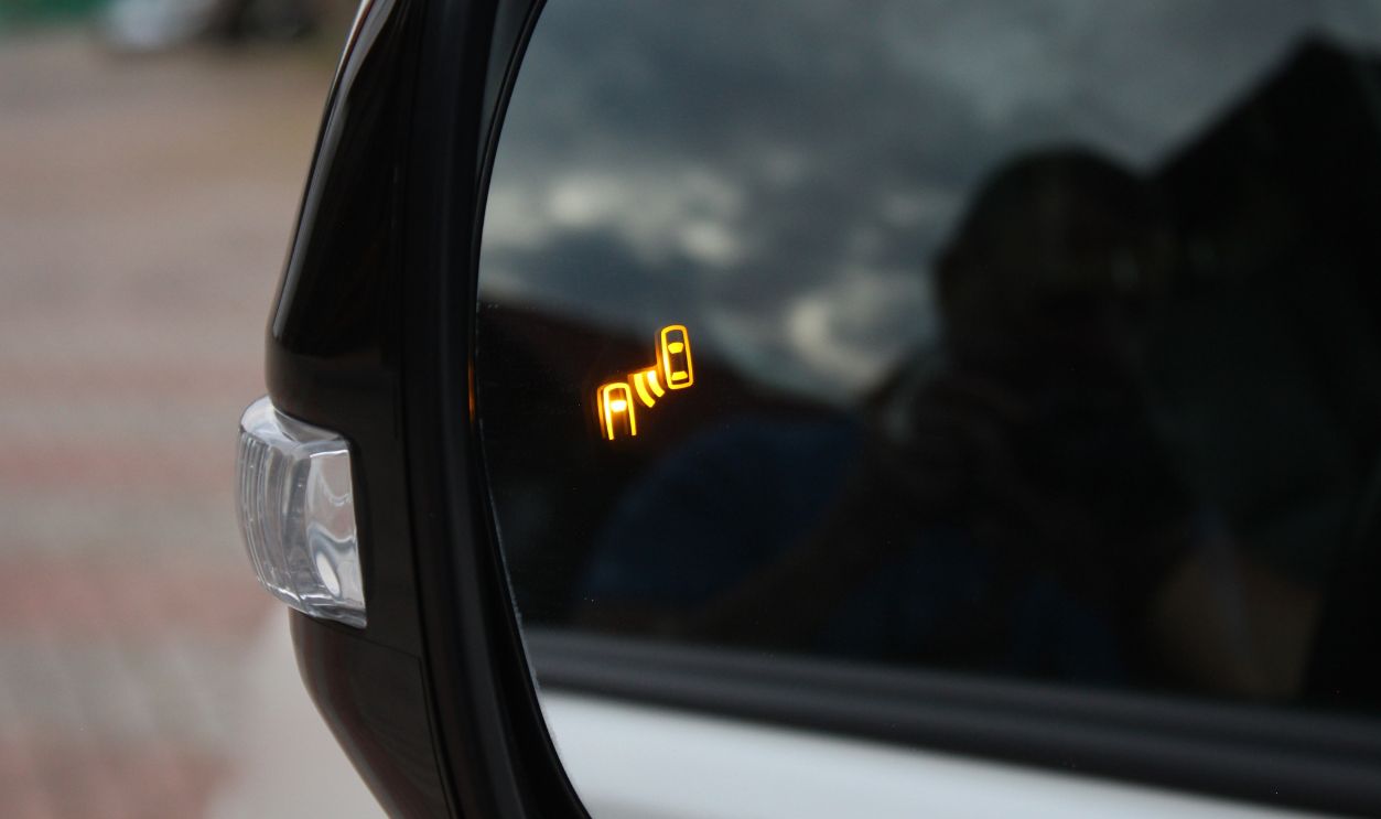 Car side mirror with illuminated orange blind spot monitoring warning indicator, glossy black frame and clear turn signal light on modern vehicle exterior.