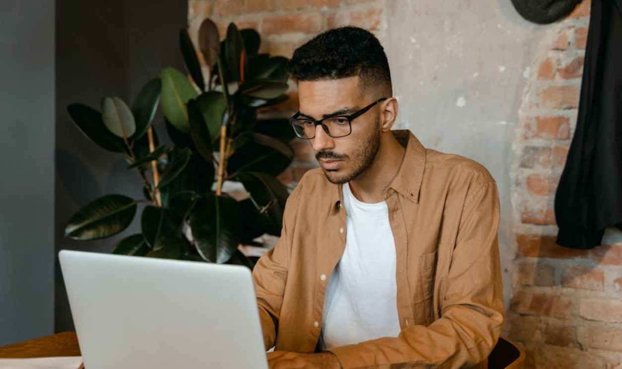 A man in glasses working on a laptop in a cozy cafe with brick walls and plants.