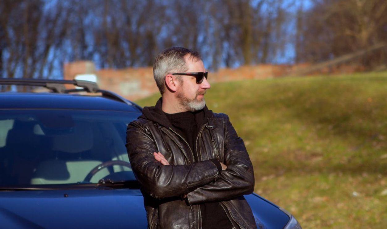 Adult man with beard posing confidently outdoors in a leather jacket and sunglasses, leaning on a car