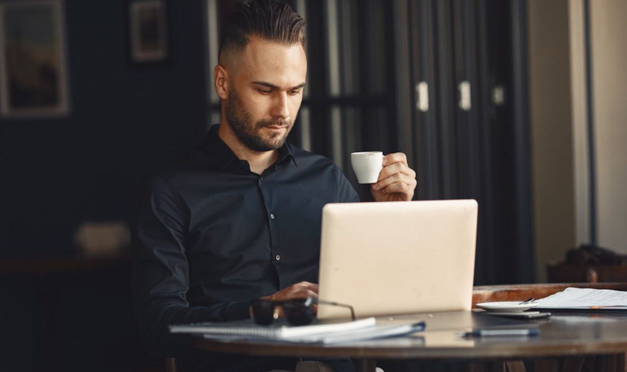 Adult man in a black shirt focused on his laptop, holding a coffee cup while working remotely indoors