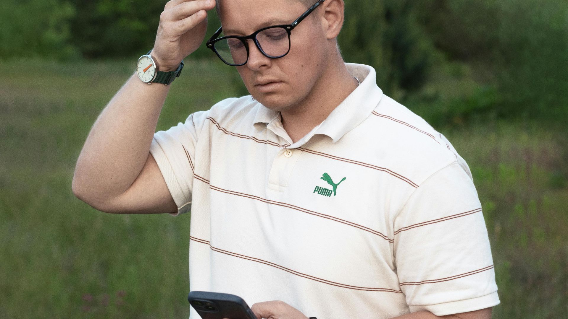 A young man in casual wear checking his smartphone in a natural outdoor setting, looking thoughtful.